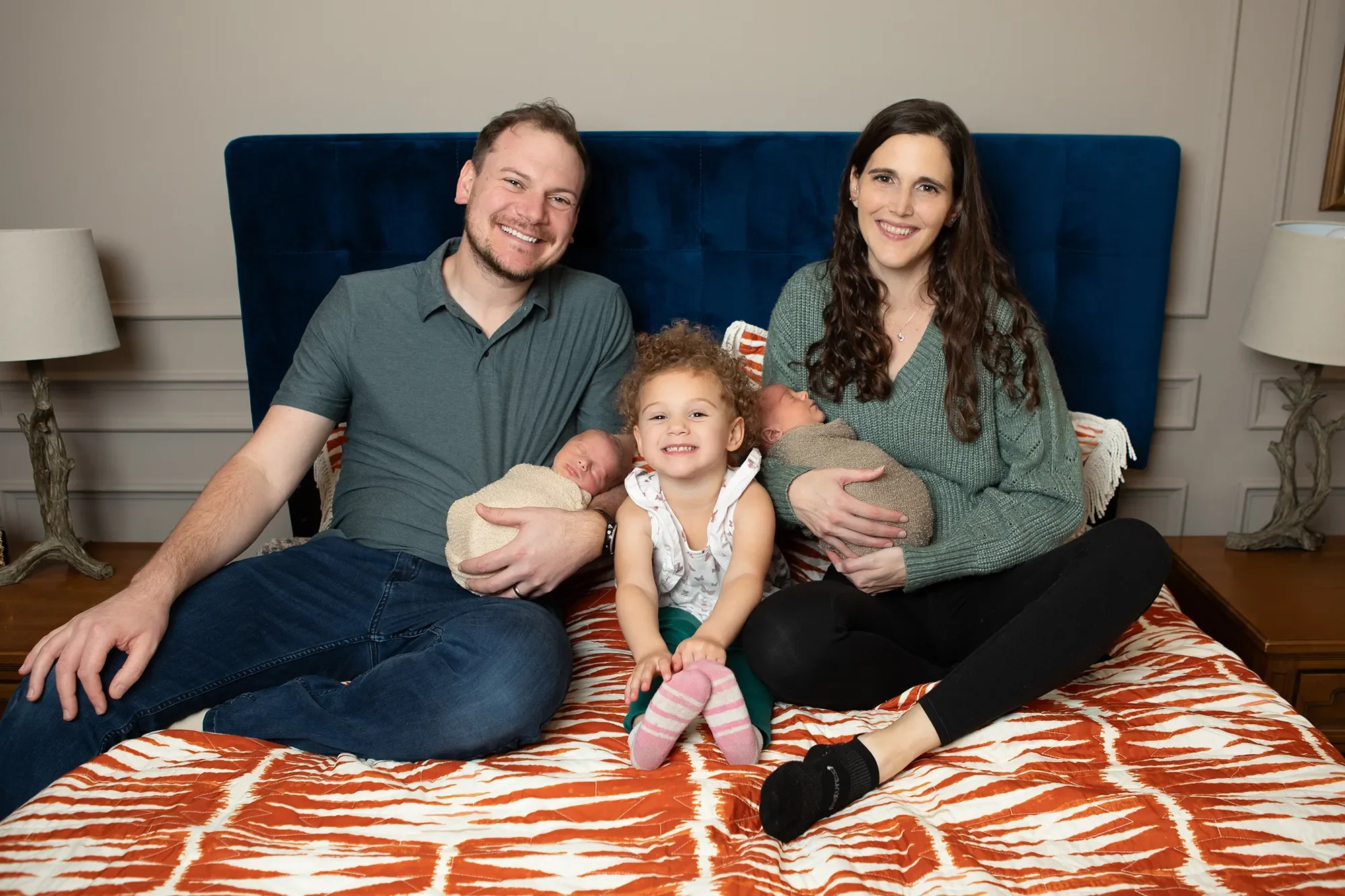 Photo of a mom and dad holding their newborn twins on their bed with their younger daughter during in home lifestyle newborn photography session in Richmond Virginia
