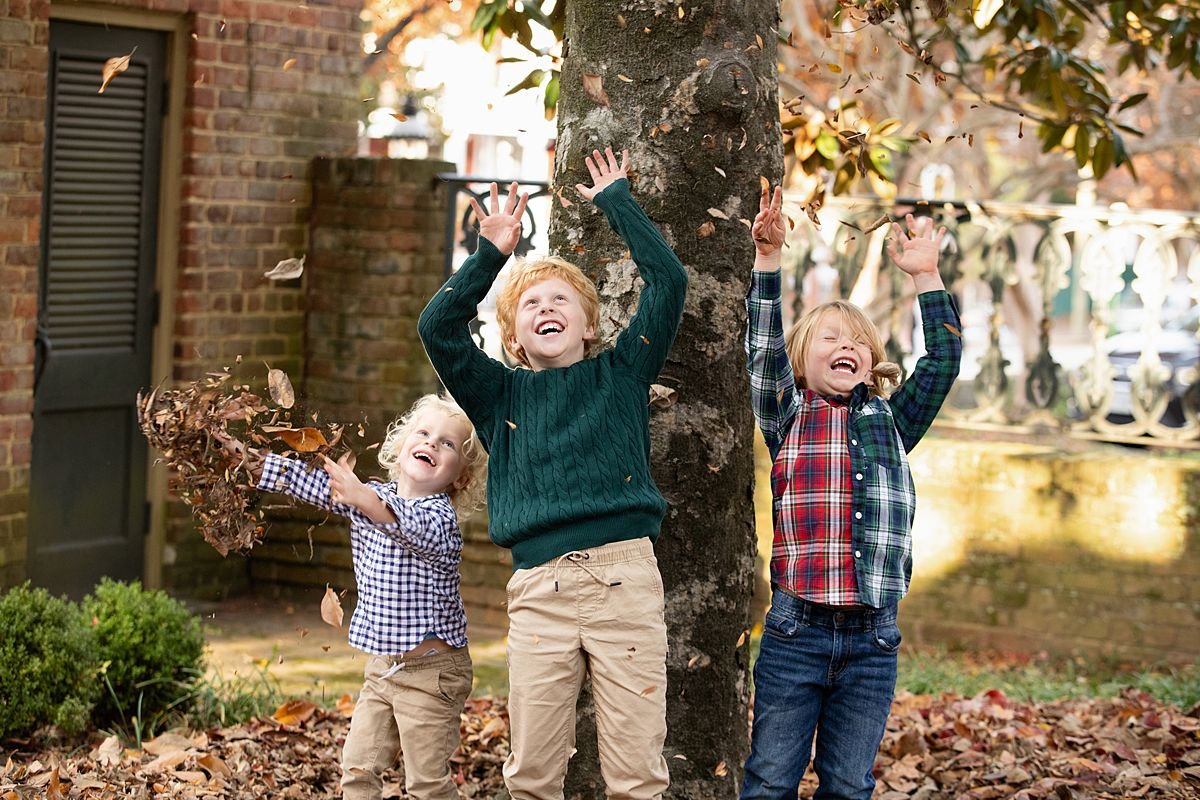 Sarah Kane Photography fall family portraits Richmond Virginia — three brothers smiling outdoors throwing fall leaves with brick garden and ornate iron gate background