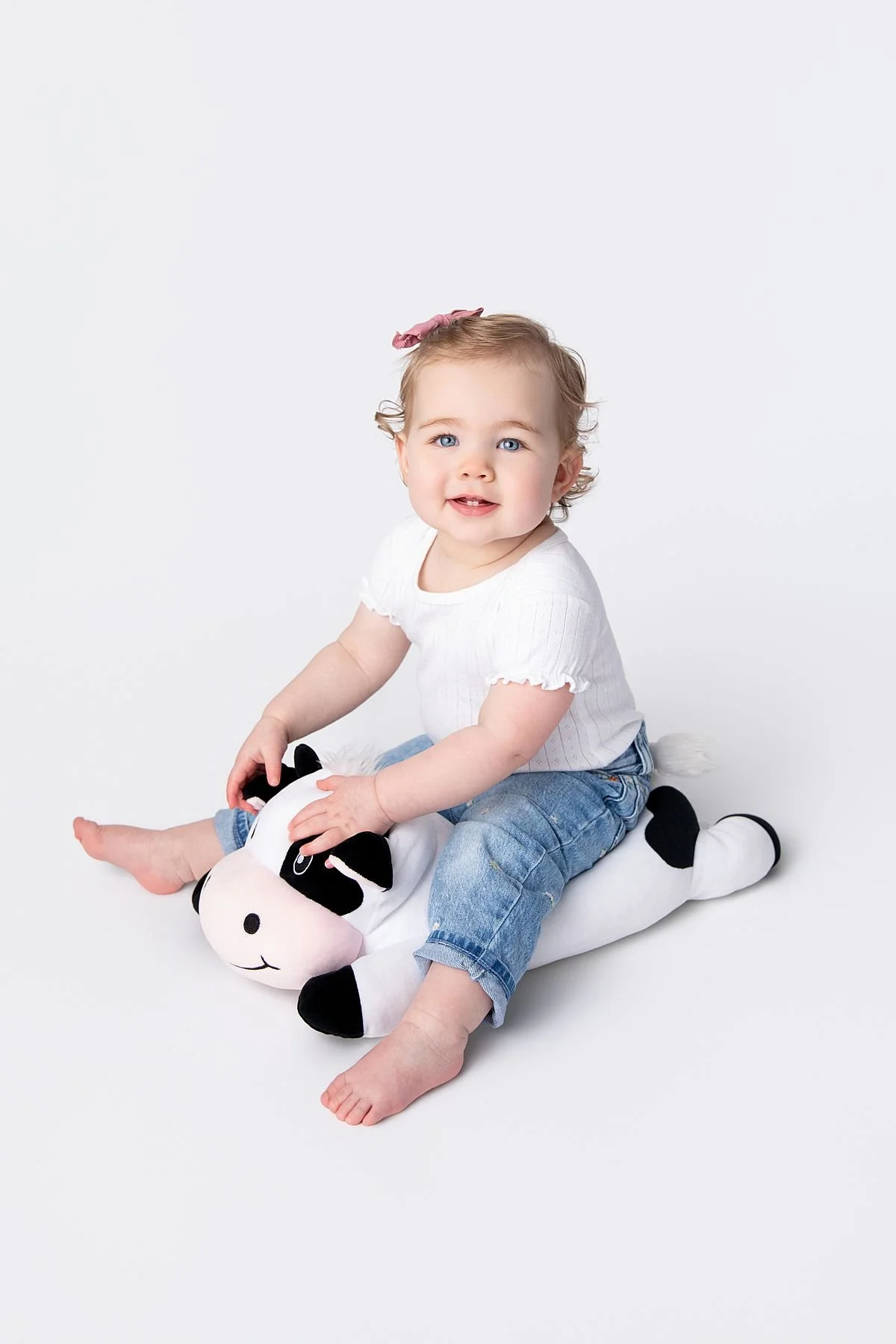 Smiling baby girl sitting on a stuffed cow in a white shirt and blue jeans, Sarah Kane Photography