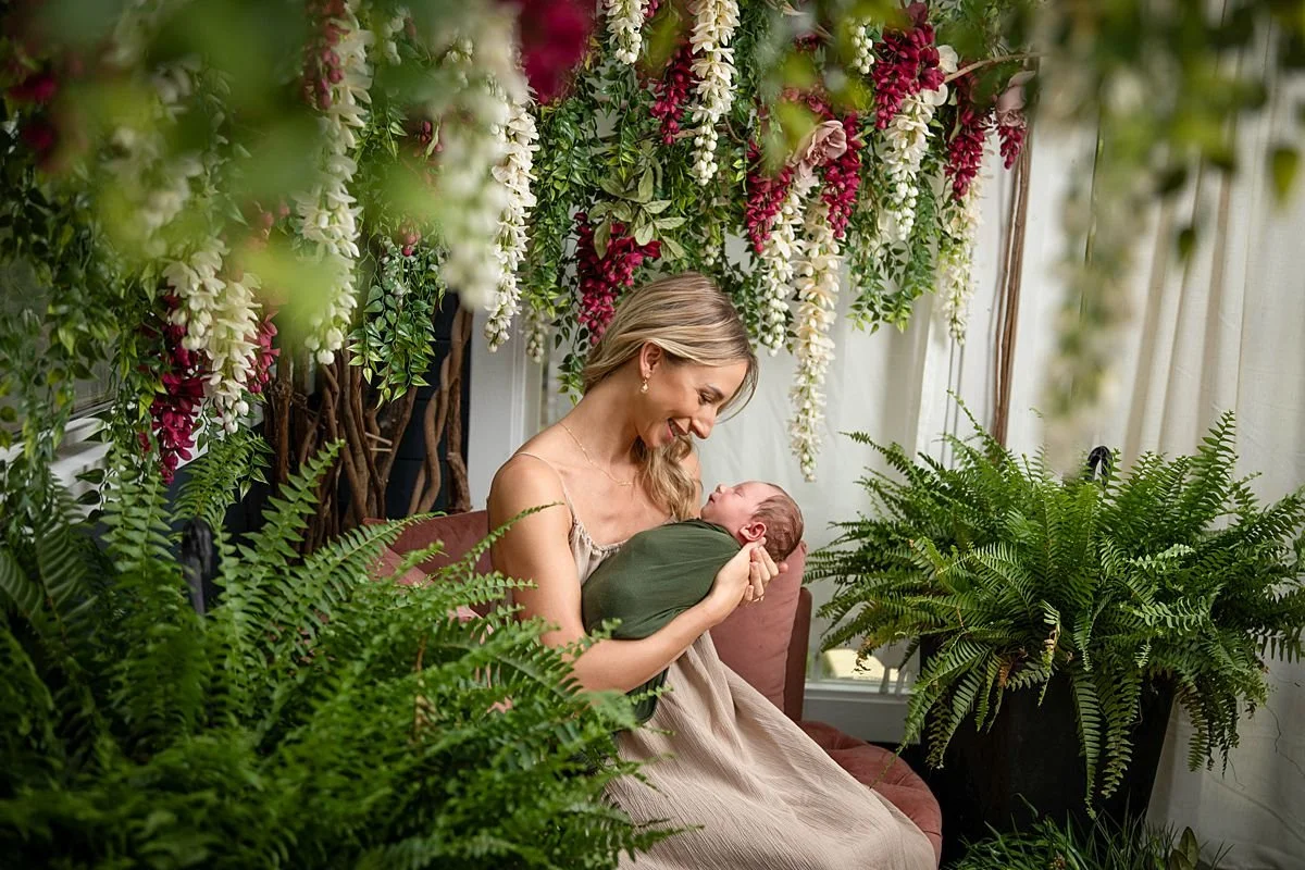 Intimate newborn portrait of mother holder her swaddled baby surrounded by cascading white and burgundy wisteria, lush ferns and greenery in a charming cottage. Newborn photographer in Chesterfield Virginia, newborn portraits near Richmond VA