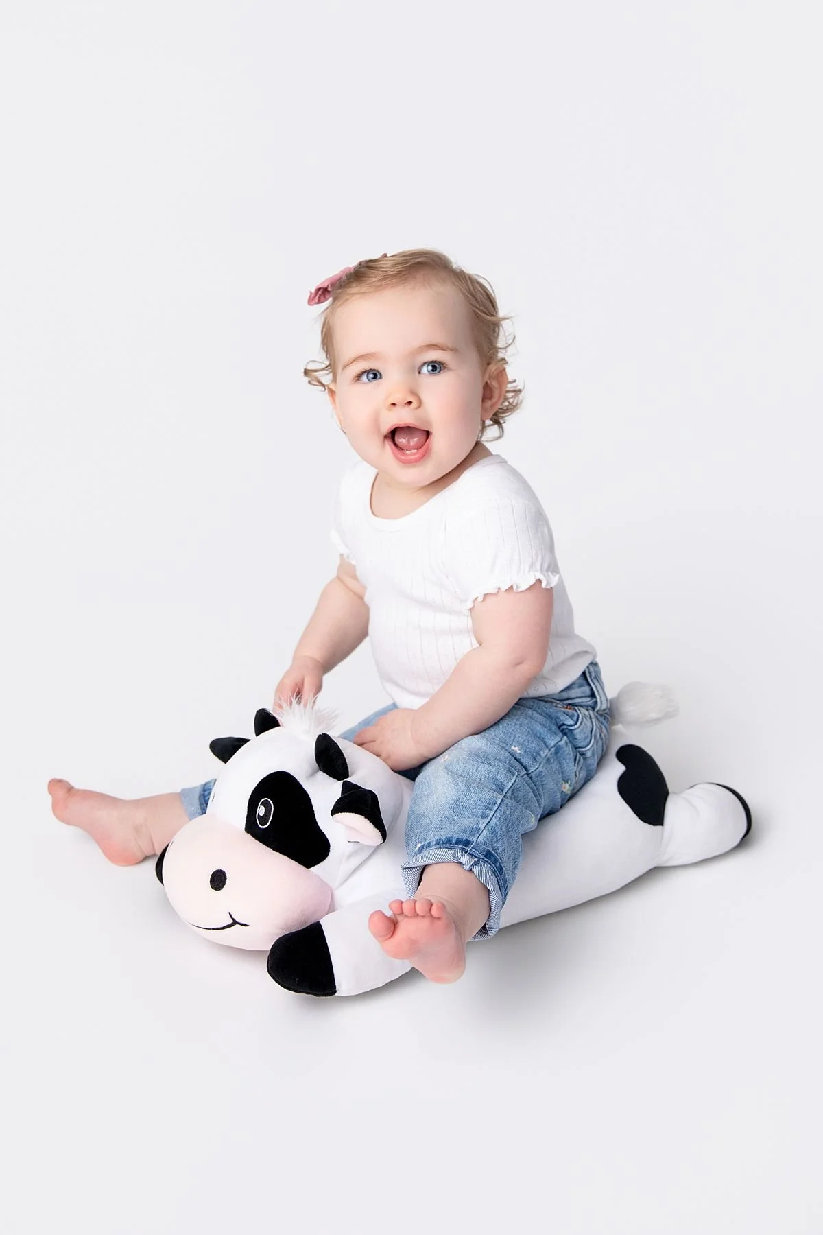 Happy baby girl sitting on a stuffed cow in a white shirt and blue jeans, Sarah Kane Photography
