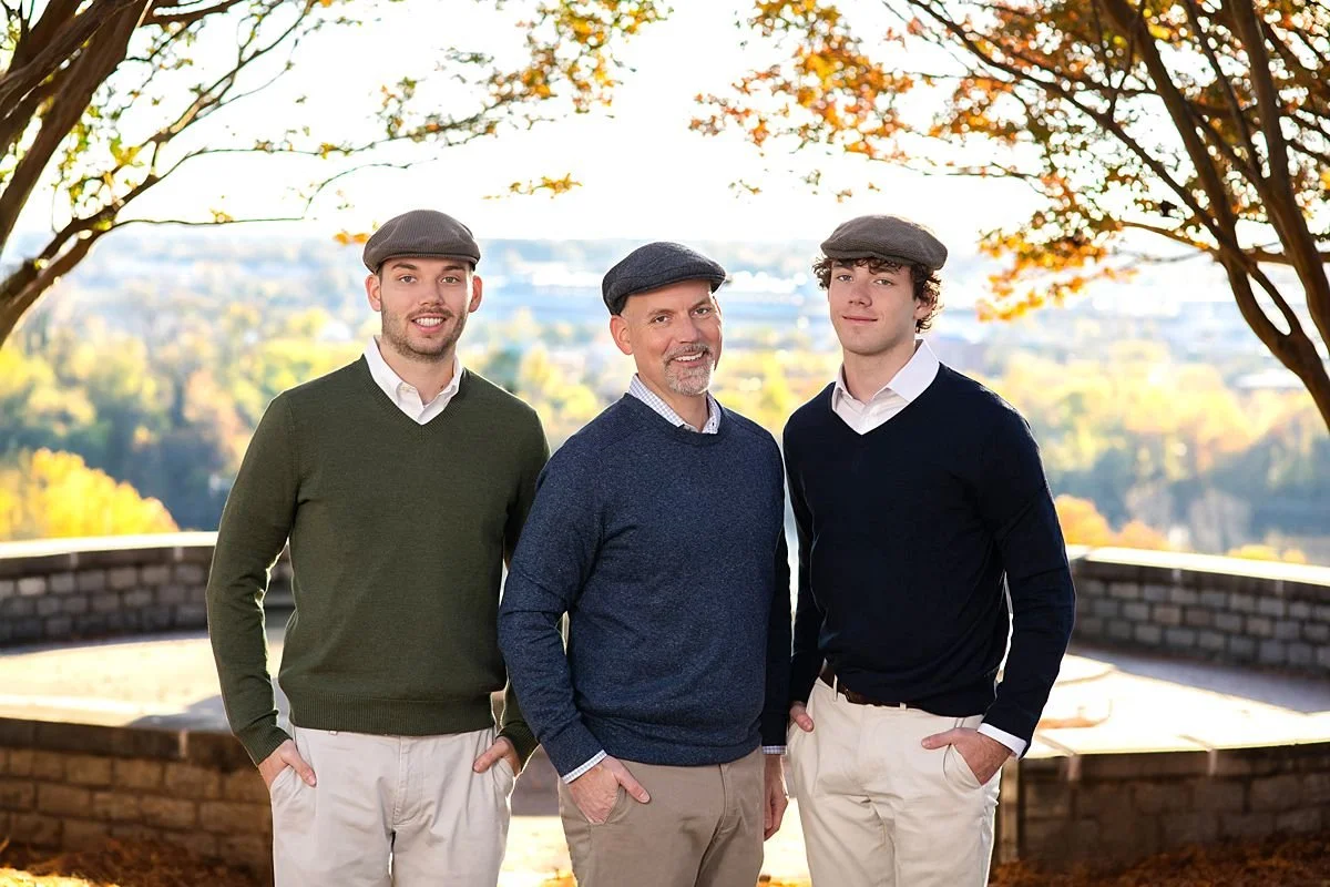 Photo of a father between his two grown sons. All wearing matching caps in front of a fountain beneath a canopy  of fall trees at Libby Hill in Richmond Virginia
