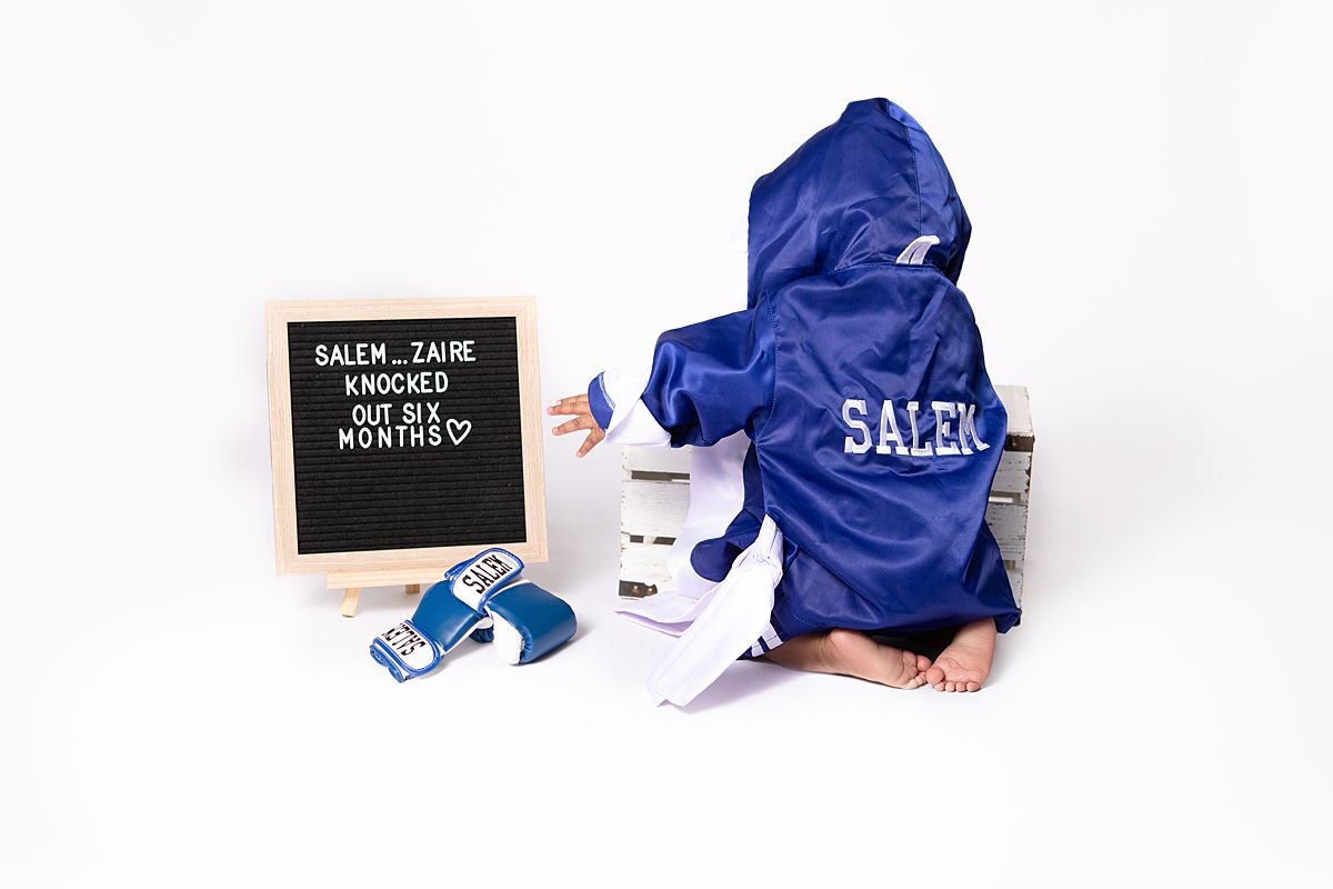 Baby in personalized boxing robe beside a letterboard reading "Knocked out six months" by Sarah Kane Photography, Chesterfield VA