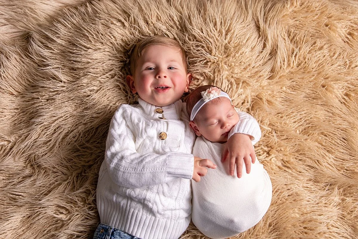 Smiling toddler boy with arm around sleeping newborn sister on fluffy rug, Sarah Kane Photography