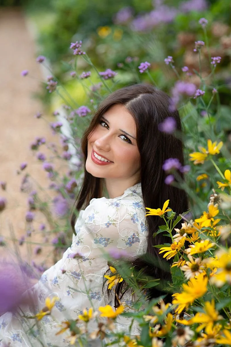 Smiling high school senior in a white floral dress surrounded by purple verbena and yellow wildflowers, Sarah Kane Photography, Maymont Park Richmond Virginia