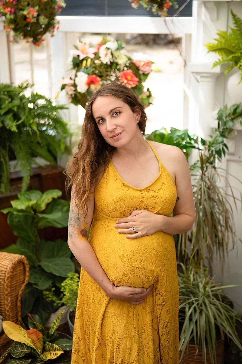 A maternity portrait of a woman with long hair, in a gold yellow dress holding her belly, with natural light, plants and flowers near Richmond Virginia. Chesterfield Photographer.