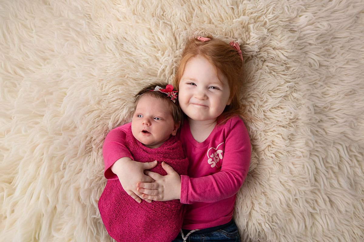 Big sister holding newborn sister on cream fur rug, Sarah Kane Photography
