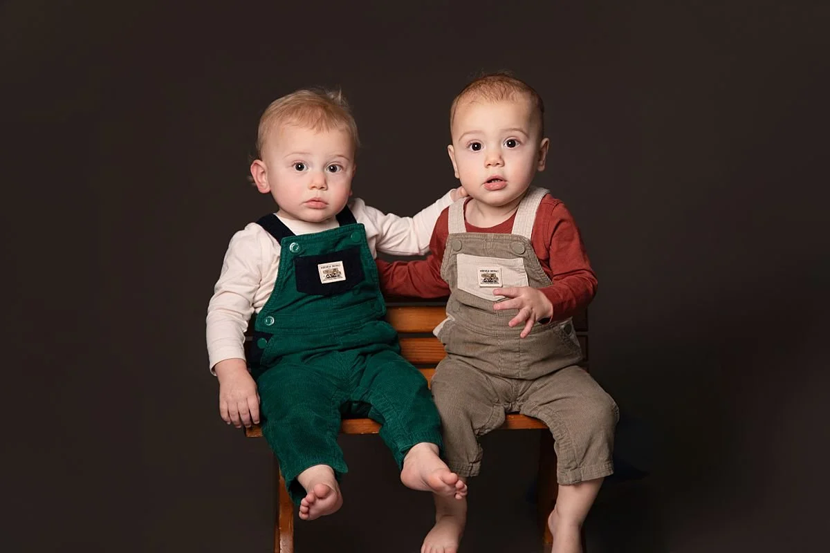 Twin one year olds sitting together on a bench looking at the camera by Sarah Kane Photography