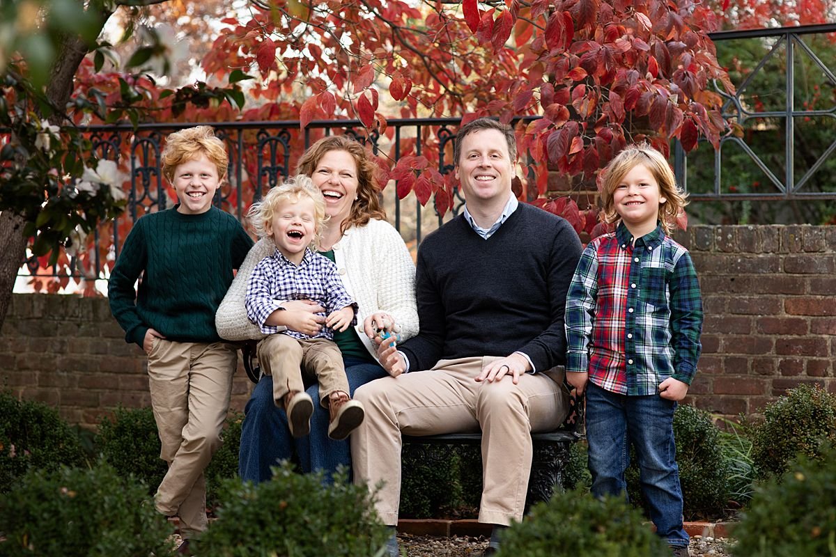 Sarah Kane Photography fall family portraits Richmond Virginia — parents and three young boys laughing outdoors with red autumn leaves and brick garden setting