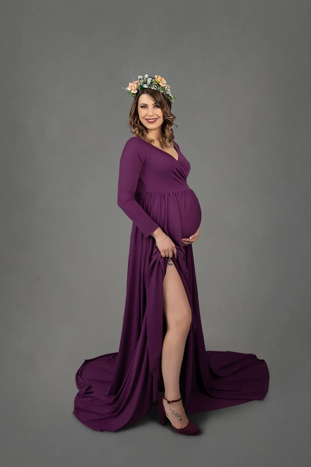 Studio Maternity Photographer in Chesterfield Virginia Just Outside of Williamsburg VA, Smiling pregnant woman holding her belly in a plum floor length dress with a leg slit