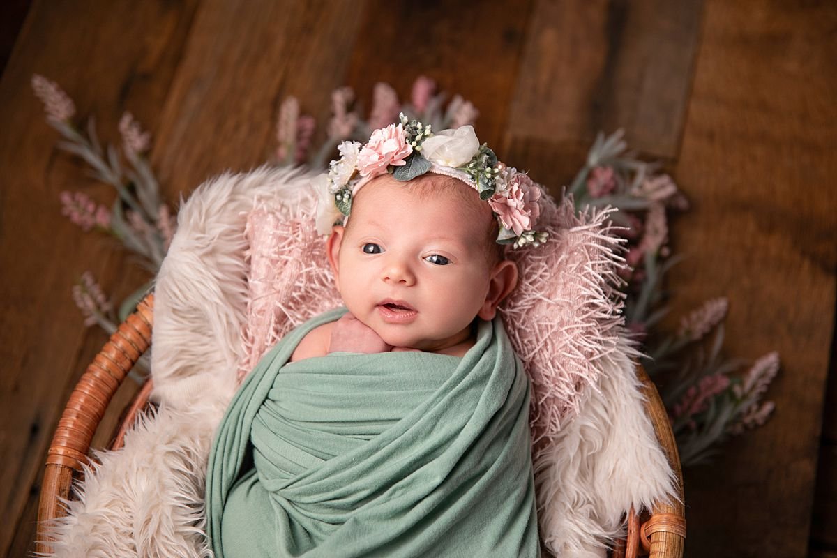 Awake newborn girl in sage green swaddle and pink floral headband in wicker basket, Sarah Kane Photography