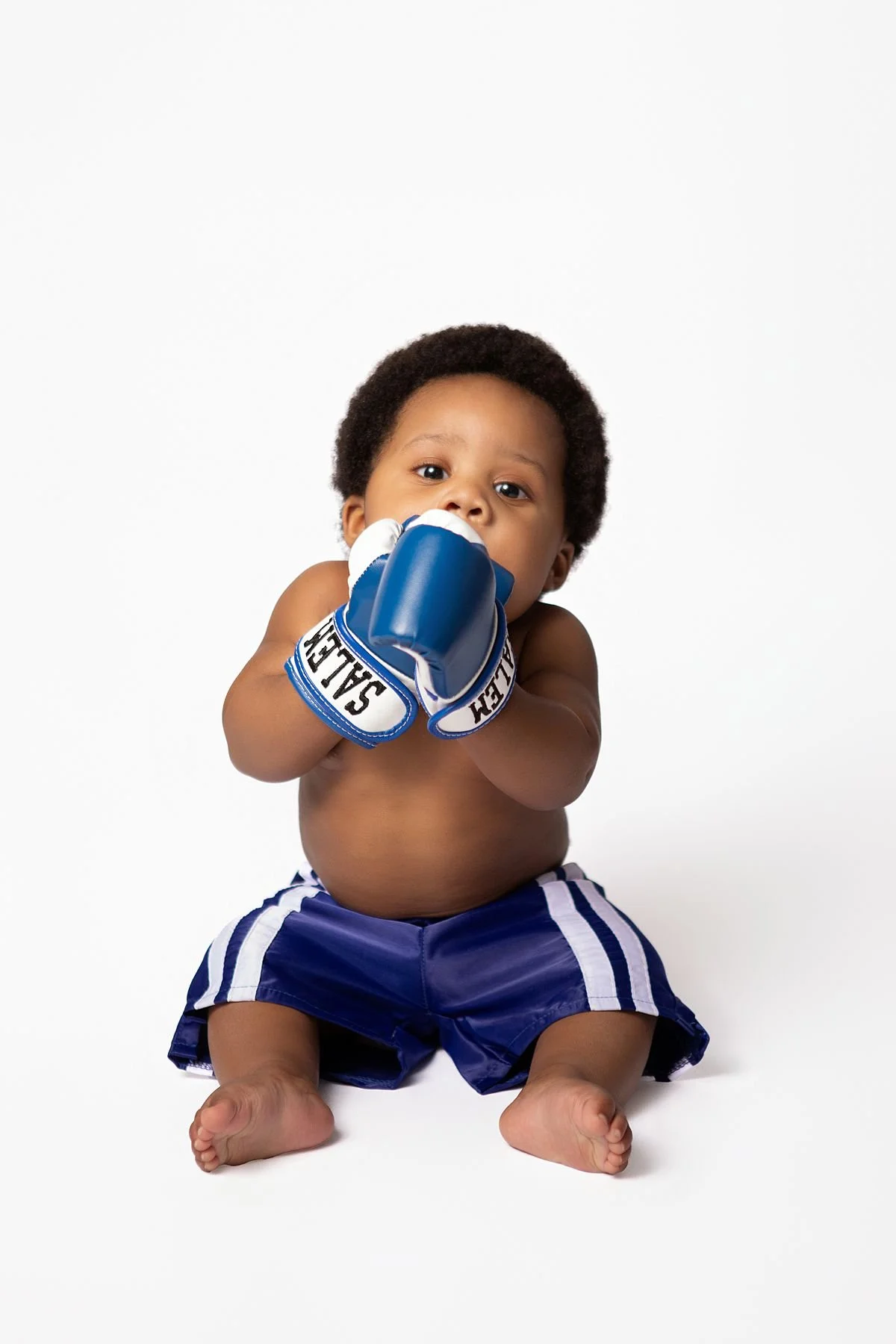 Baby Salem in boxing shorts holding personalized boxing gloves to his mouth, 6 month milestone by Sarah Kane Photography