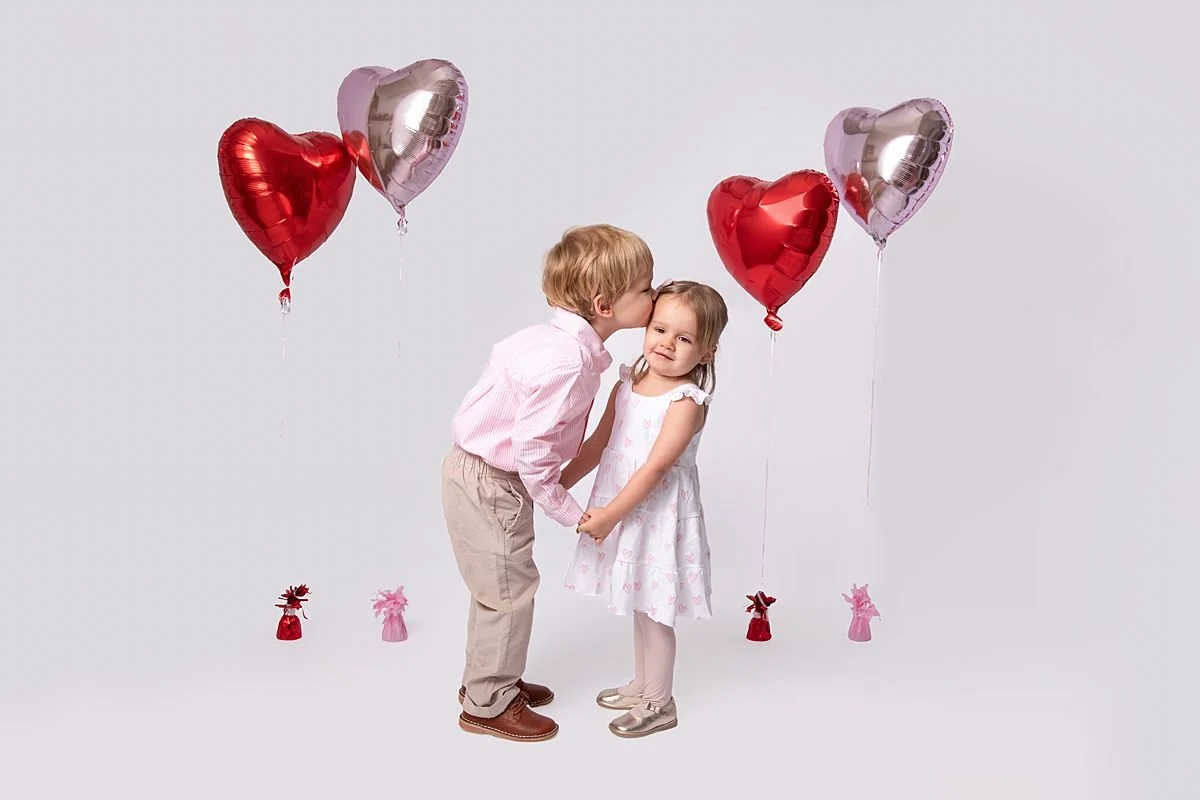Brother kissing his little sisters head while holding hands in front of valentines balloons photographed by Sarah Kane Photography near Richmond VA