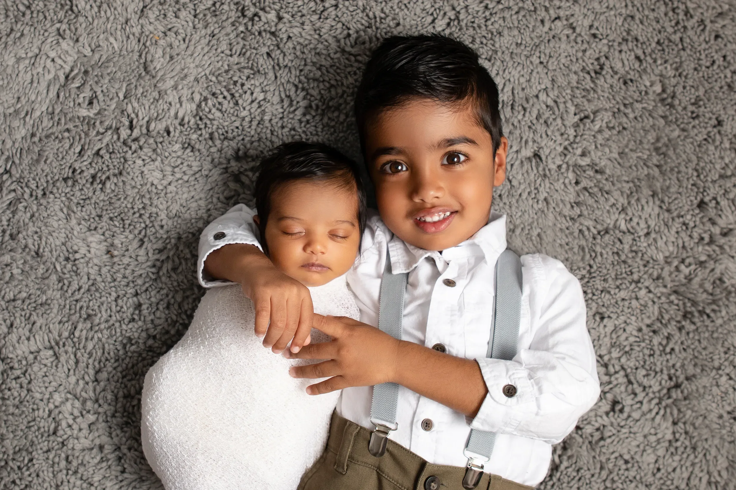Photo of 4 year old big brother holding his newborn brother wrapped in a white swaddle on a grey rug for newborn portraits in Chesterfield Virginia