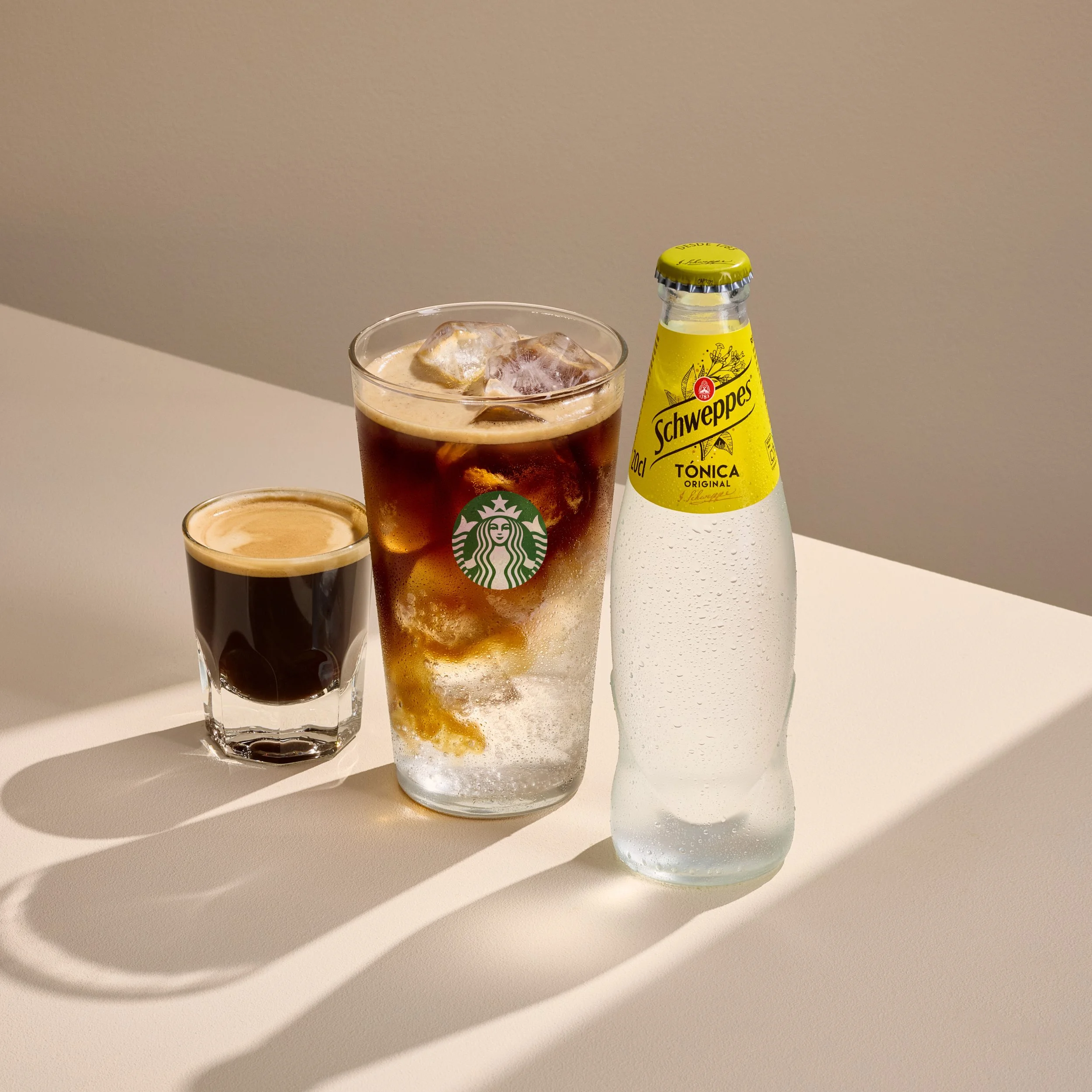 Espresso Tonic de Starbucks sobre hielo junto a una botella de Schweppes Tónica Original y un chupito de espresso, sobre fondo beige con luz natural.
