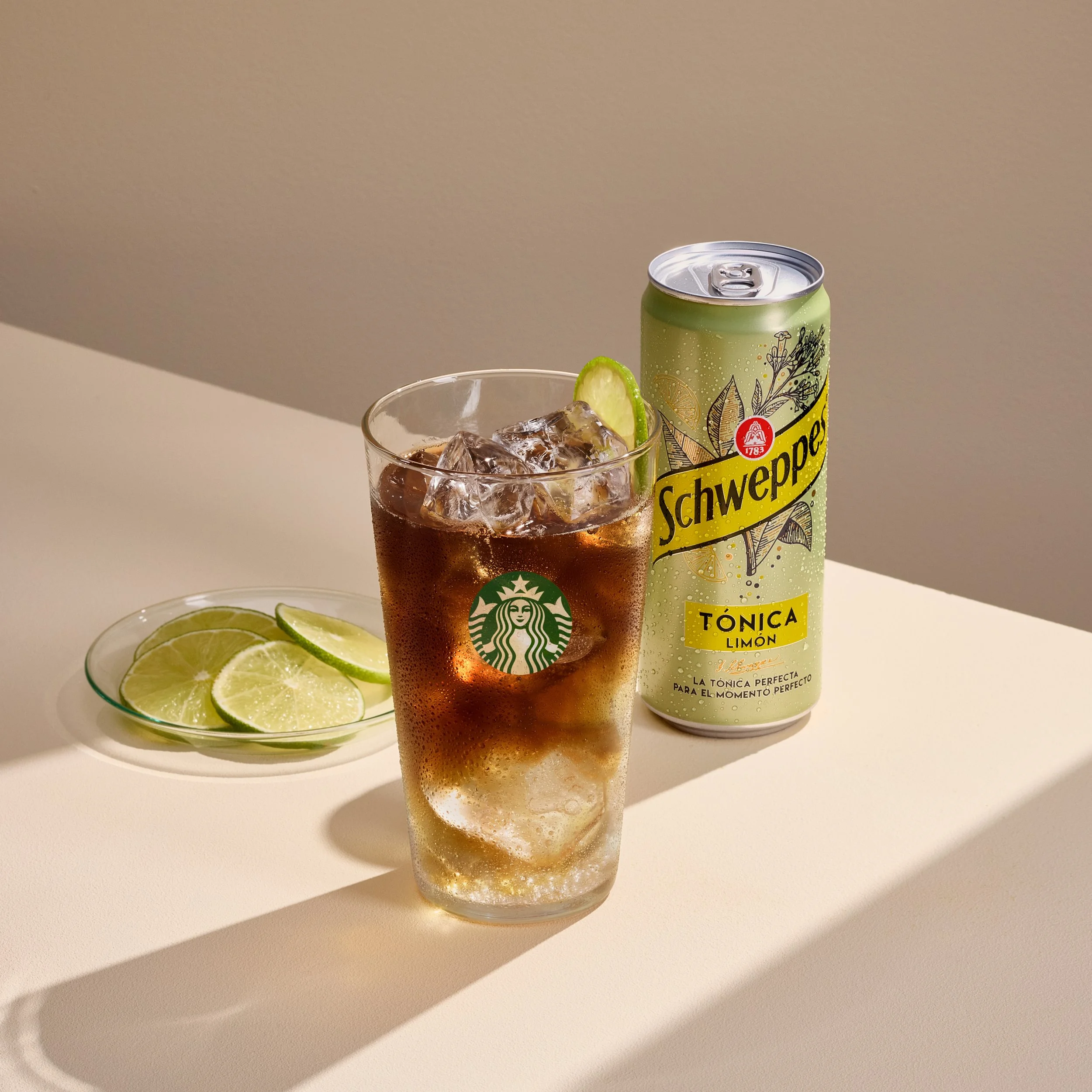 Cold Brew Tonic de Starbucks con hielo y una rodaja de lima, junto a una lata de Schweppes Tónica Limón y un platito con rodajas de lima, sobre fondo beige.