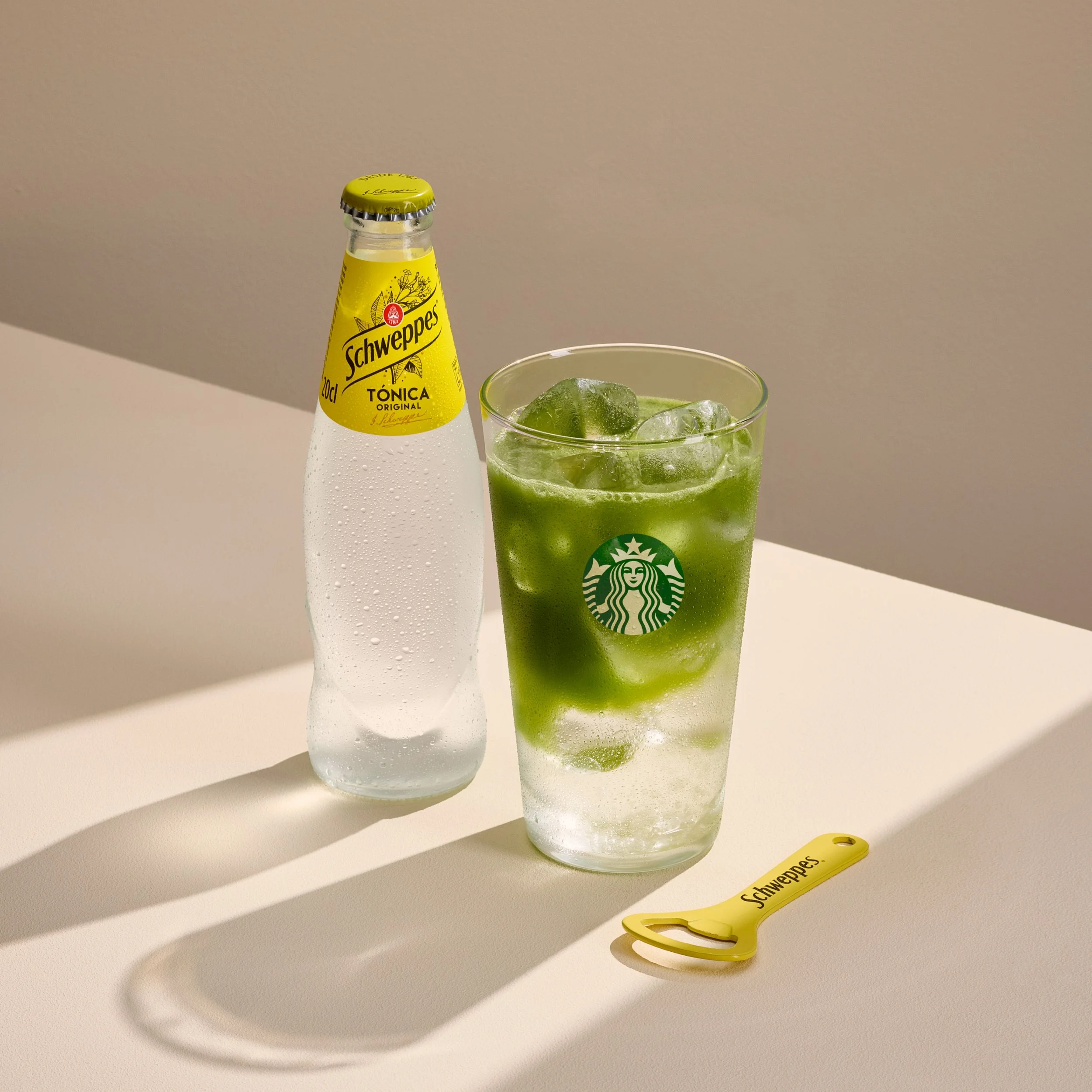 Matcha Tonic de Starbucks, bebida verde con hielo, junto a una botella de Schweppes Tónica Original y un abridor amarillo Schweppes sobre fondo beige.