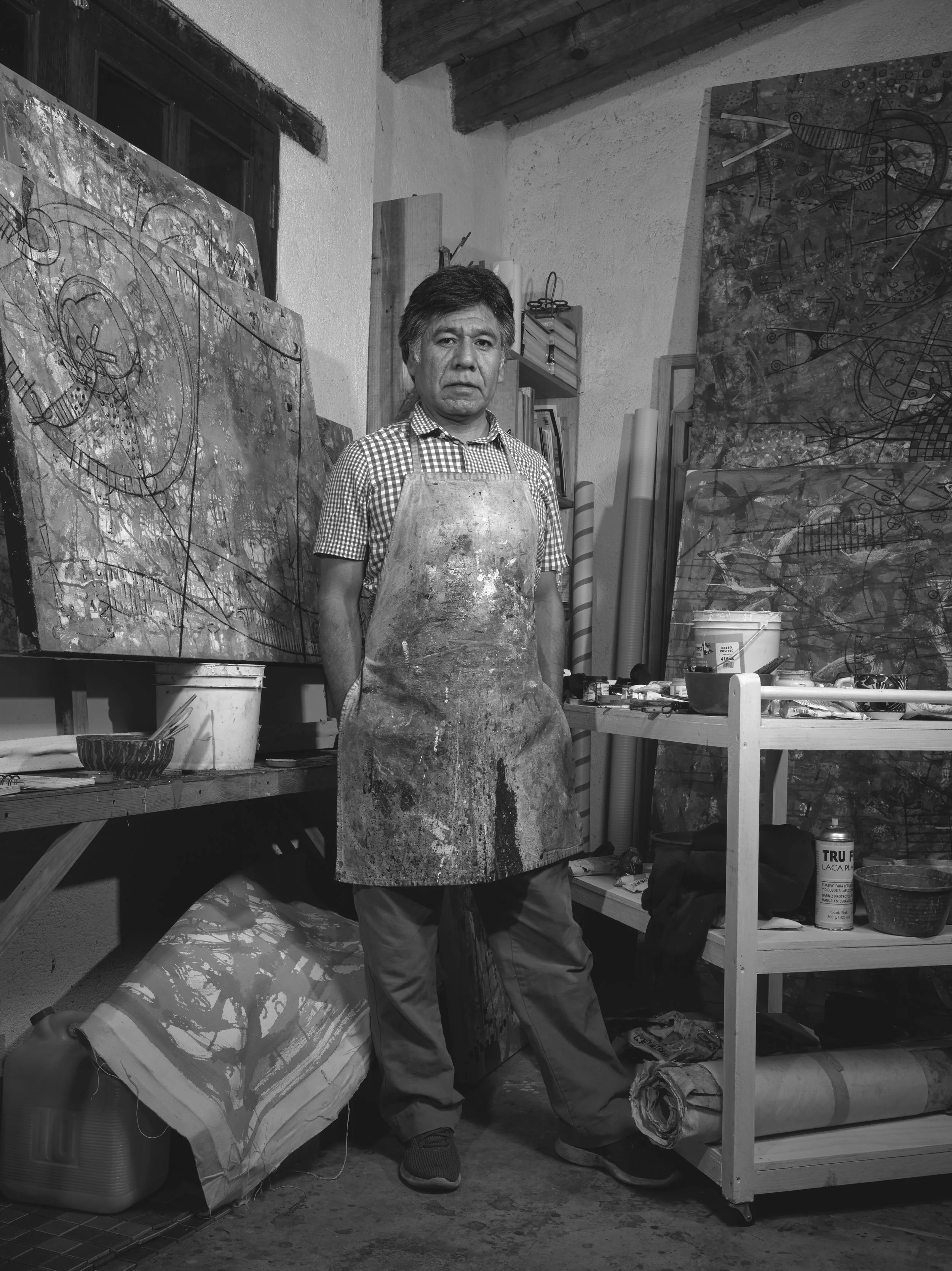 Black and white artist portrait of Fulgencio Lazo, standing surrounding by paintings and a cart with supplies, wearing a paint splattered apron.