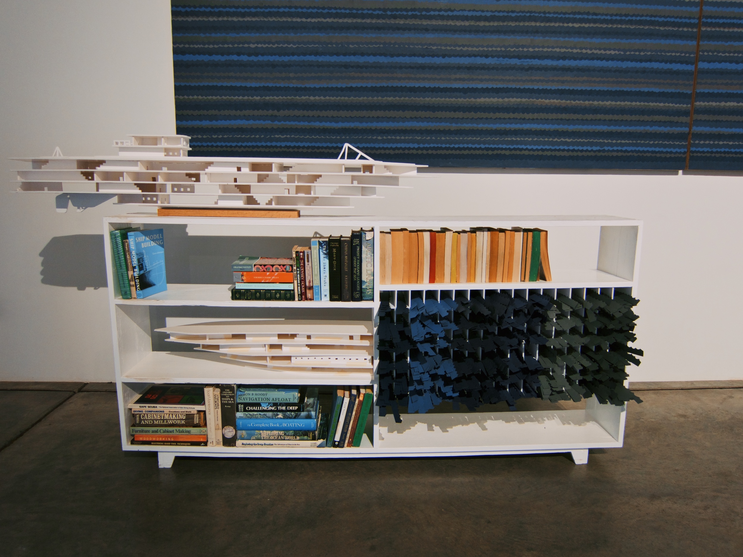  Built plywood cabinet containing books (adventure novels, woodworking manuals, scuba diving field guides, and sailing handbooks), cut paper waves, and foam ship models, sitting underneath a depiction of the sea from cut paper waves glued to plywood 