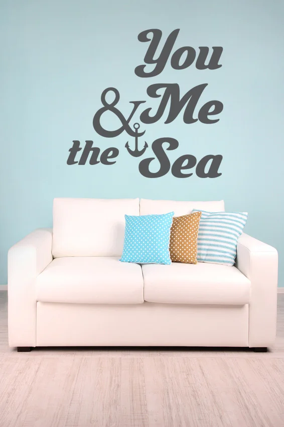 Nautical inspired wall decal quote - 'You Me & the Sea' with an anchor in dark grey on a light blue wall above a white couch with geometric throw pillows.
