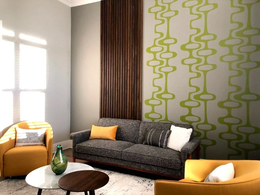 Mid Century geometric pattern wall decal by Wall Star Graphics shown in olive on a white wall above a modern grey couch and two yellow chairs.