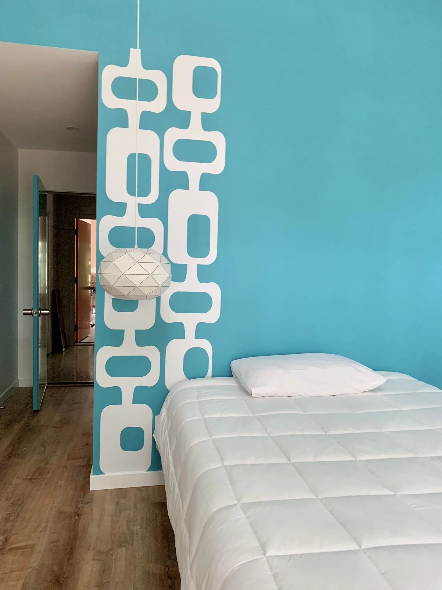 A mid century geometric pattern wall decal shown in white on a turquoise wall behind a bed in a guest bedroom.