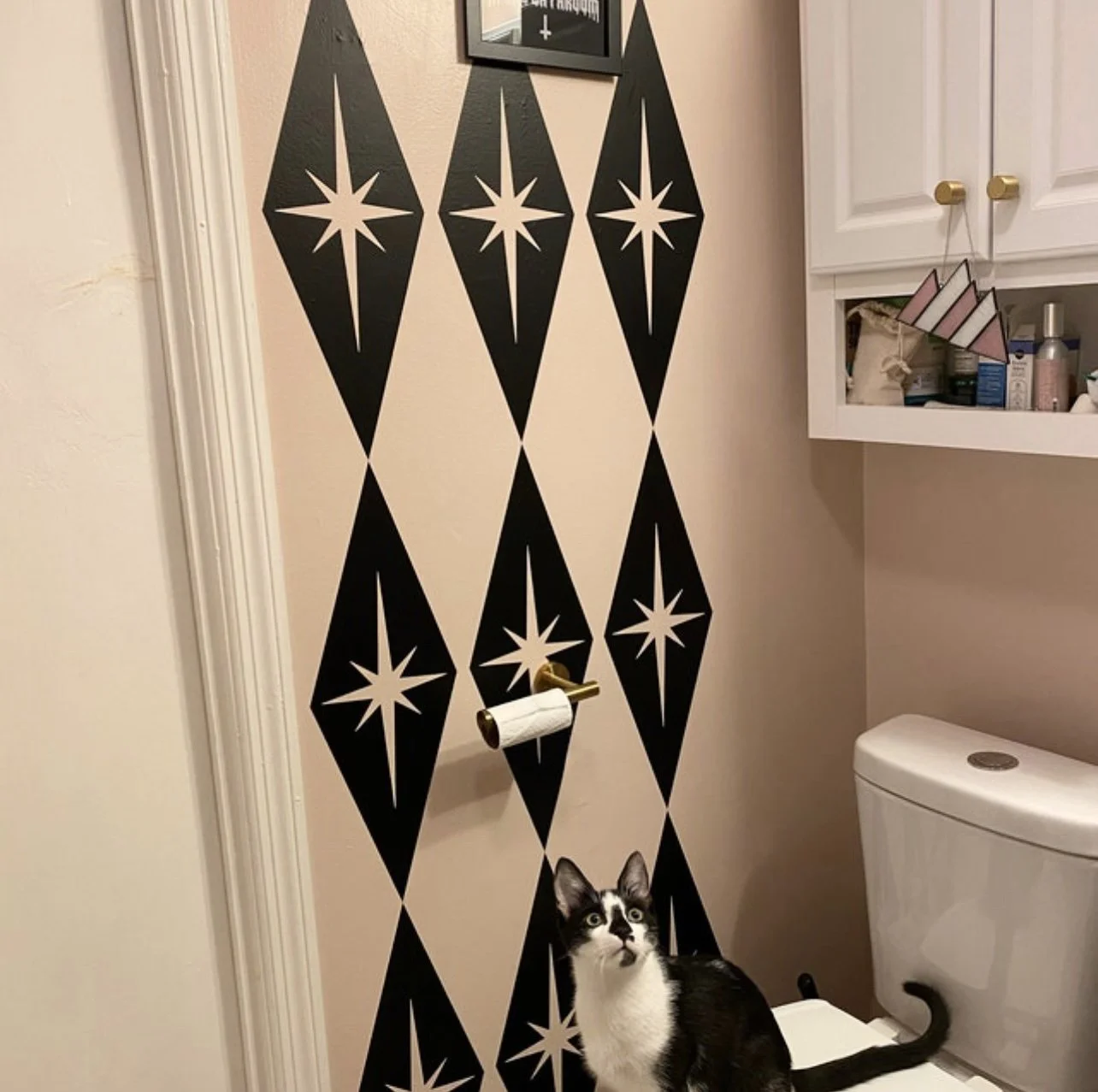 Mid century atomic style wall decals consisting of large diamond shapes with atomic stars shown in black on a light pink bathroom wall with a black and white cat sitting on the toilet.