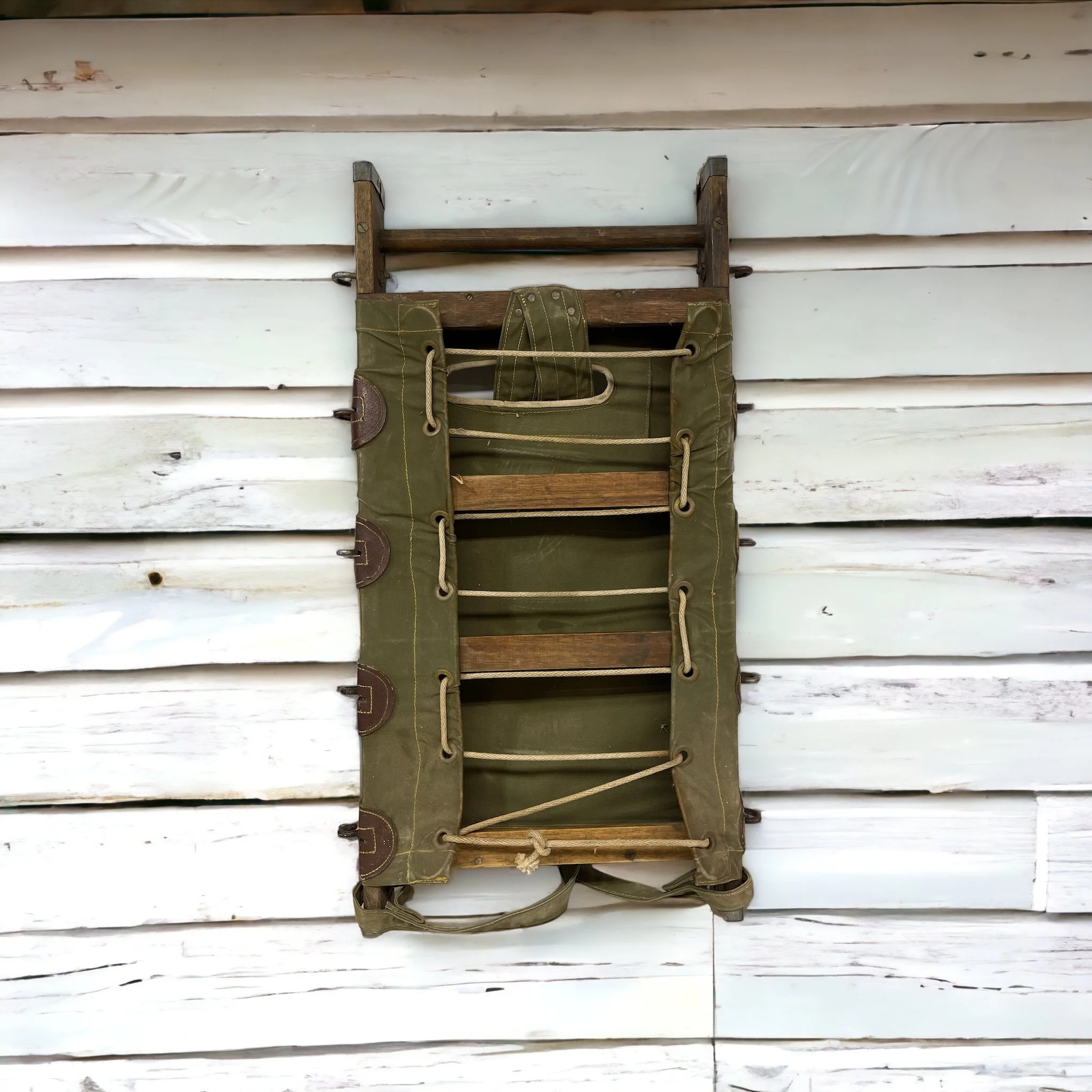 Vintage US Forest Service Wood Packboard Backpack and frame with Army green canvas, leather and metal accents with weathered wood hanging on a rustic white wooden slat wall.