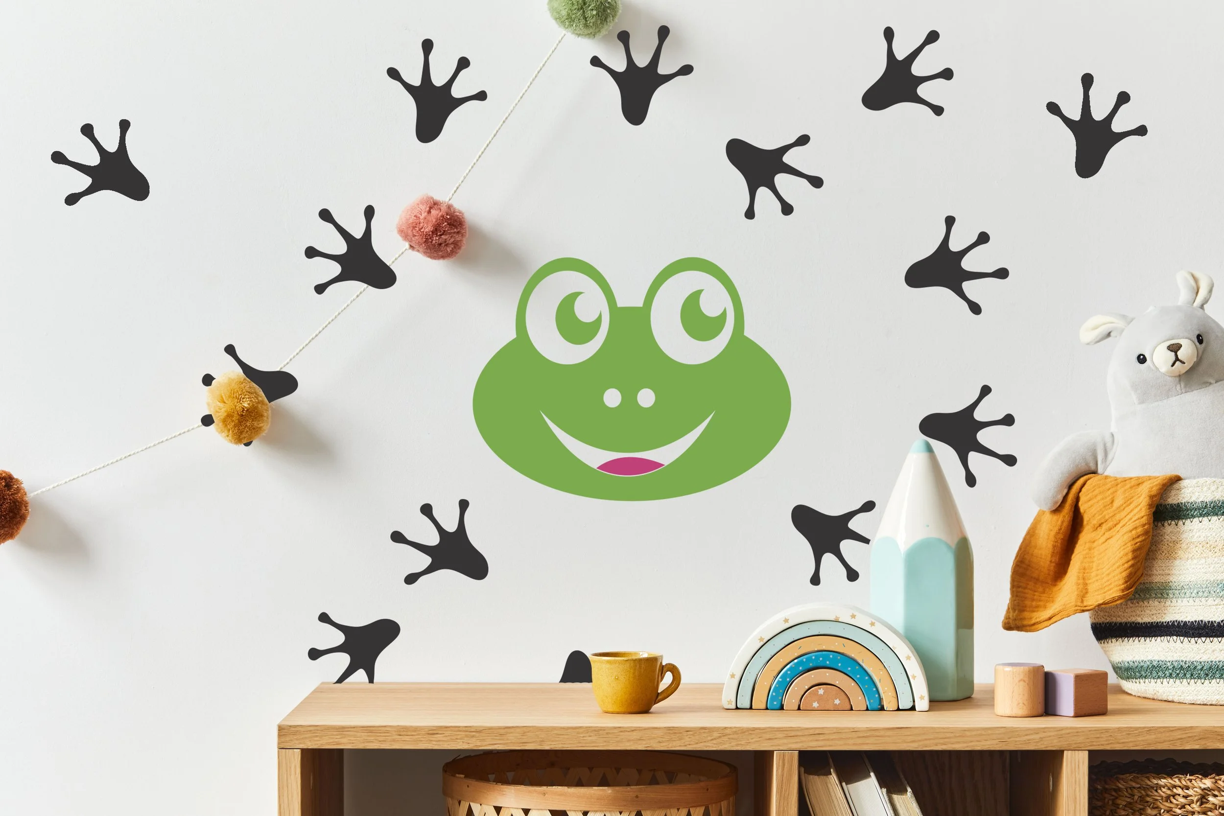 Adorable frog face and tracks wall decals styled above a light colored wooden shelf in a kids bedroom.