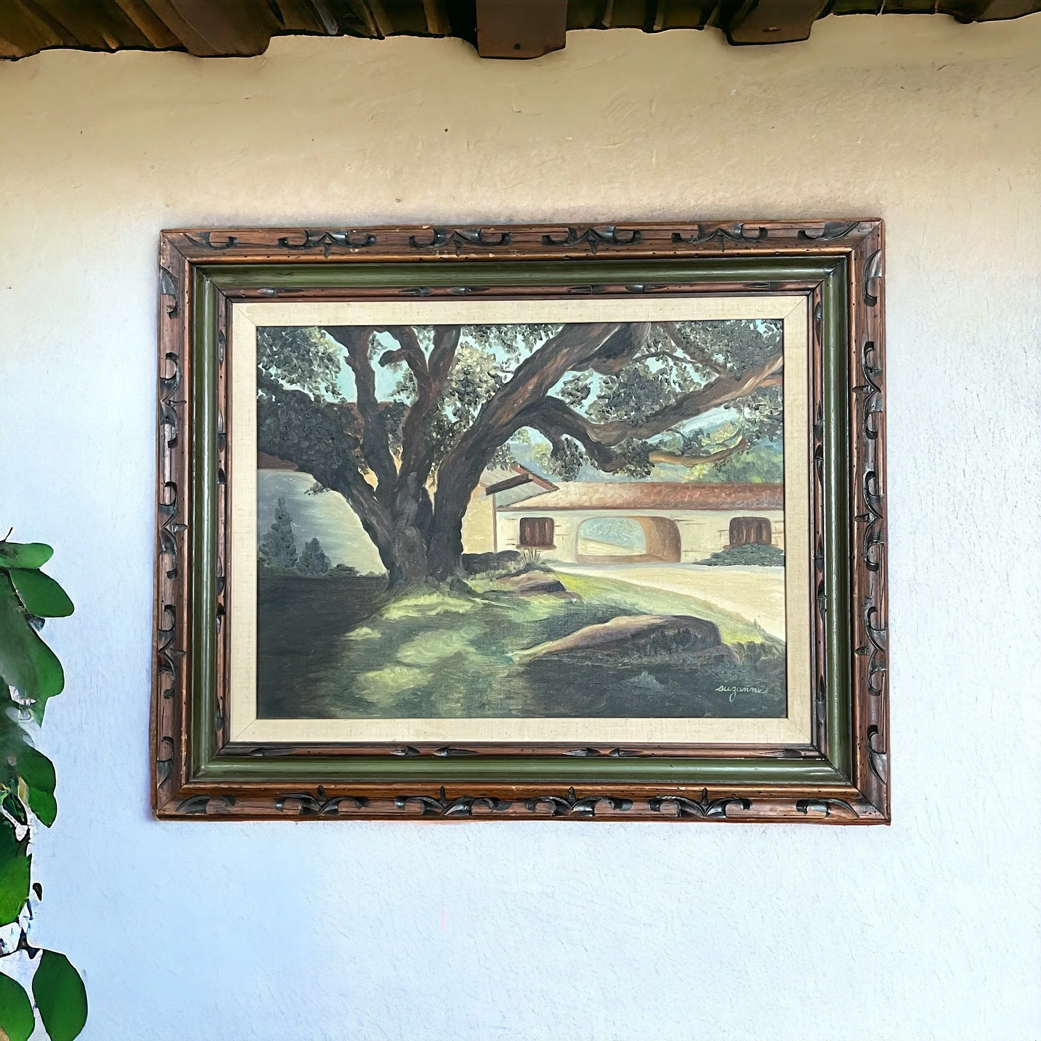 Vintage 32 x 26 inch original painting of oak tree and Spanish style home, in muted earth tone colors in a hand carved wood frame shown hanging on a stucco wall.