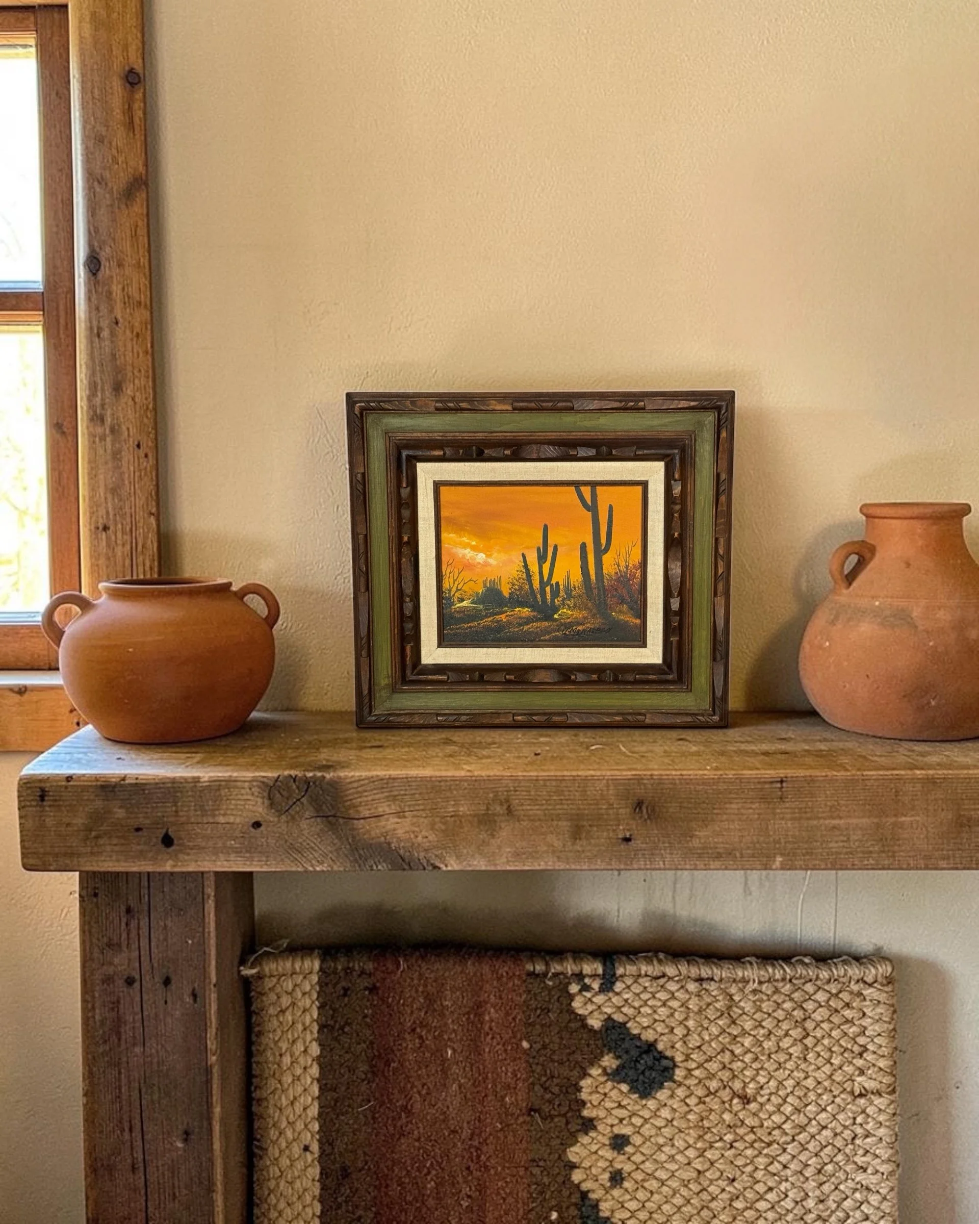 Vintage cactus landscape painting with carved framed detail styled with terracotta pots on a rustic wooden table in a southwestern style home