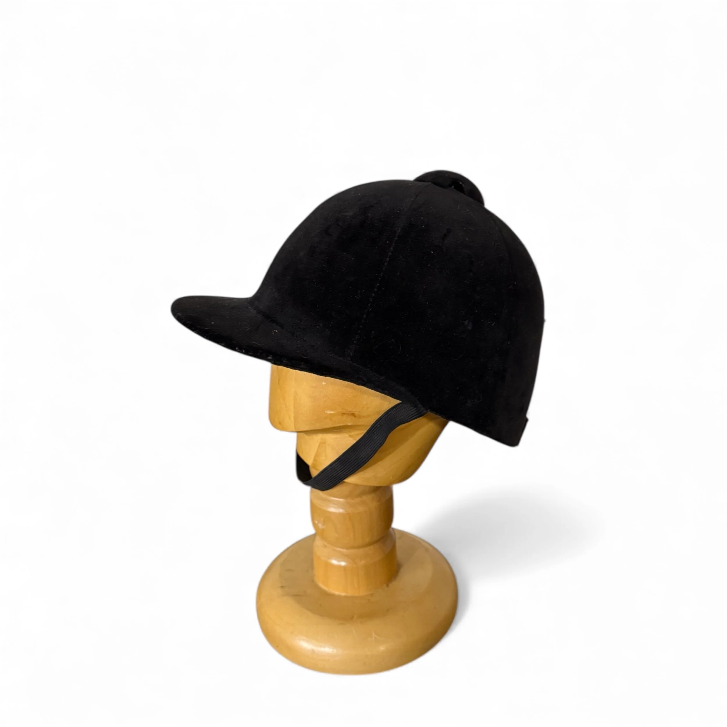 A black equestrian helmet with a rounded crown and a small brim. The helmet is displayed on a wooden head form with a chin strap. The helmet is made of a soft, velvety material.