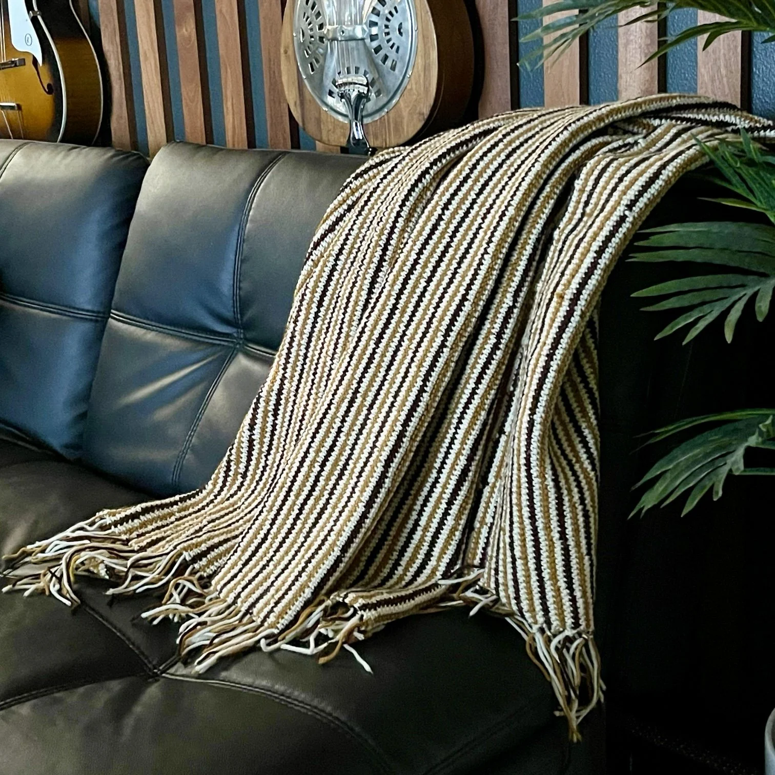 Vintage handmade crochet blanket with fringe hanging on a vintage wooden hanger. Blanket is done in a geometric line pattern on the corner of a black leather couch.