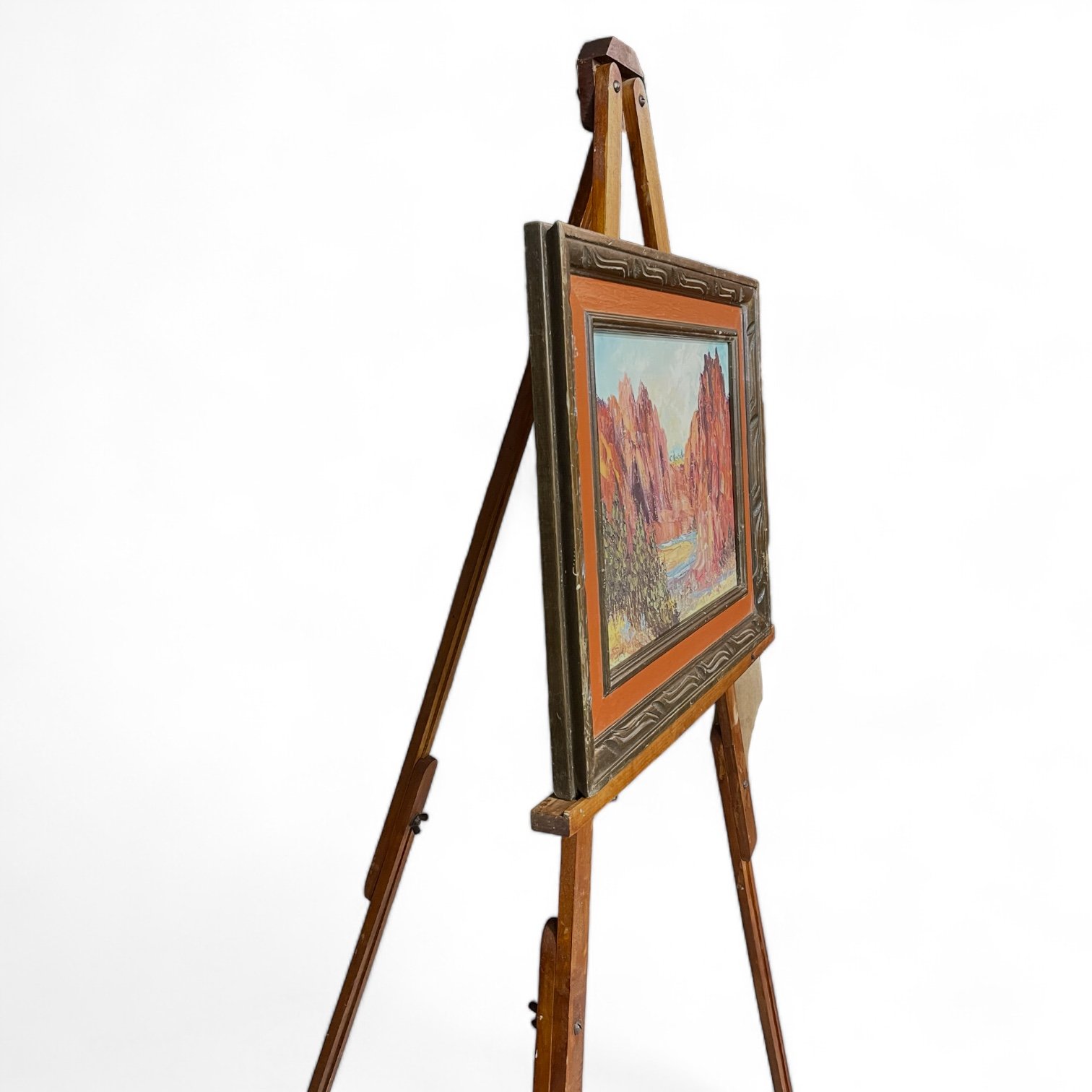 A side view of a vintage wooden adjustable floor easel displaying a framed and matted vintage oil painting of a rock canyon with a river, trees, and shrubs.