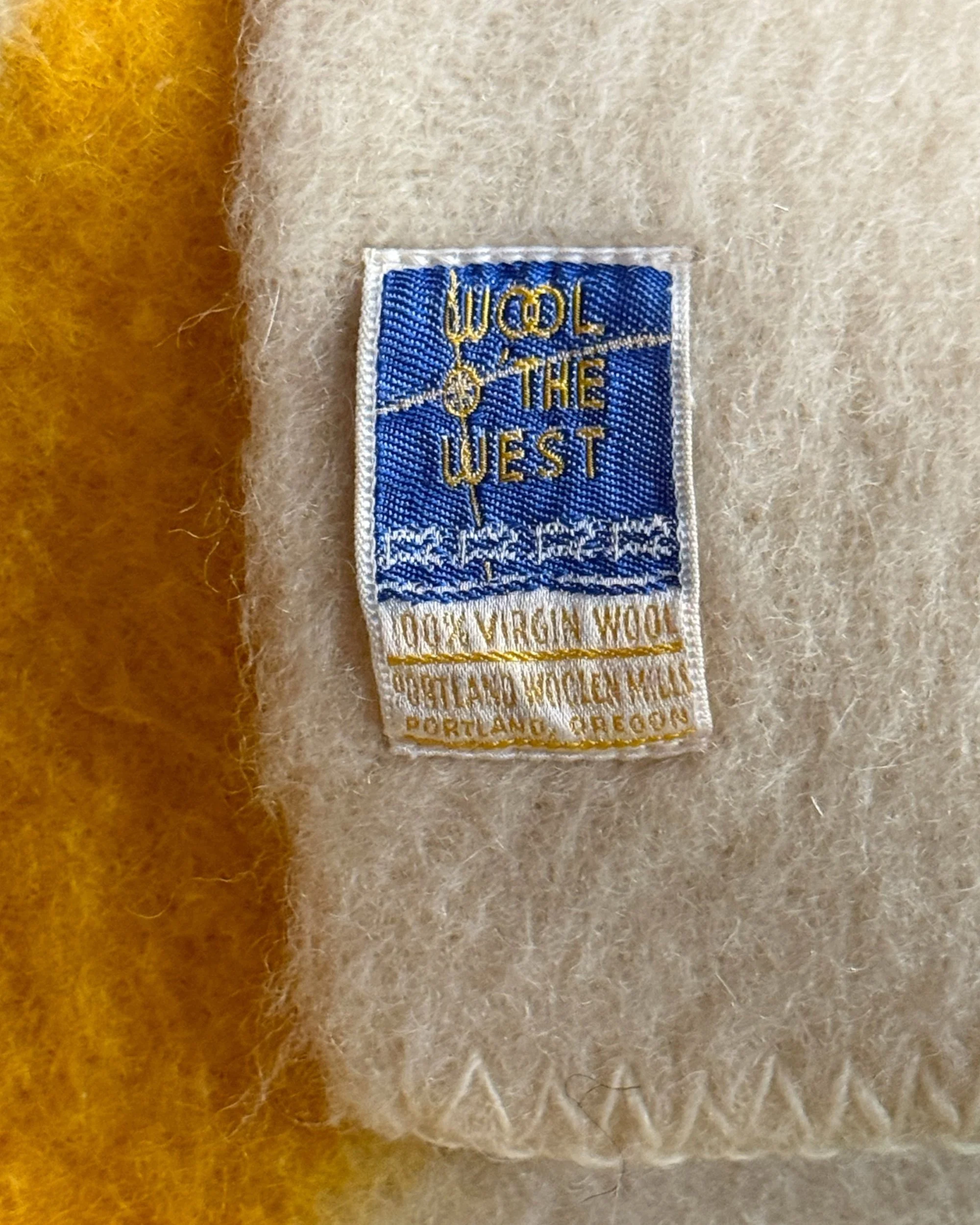 Close-up of the vintage blue and gold Wool O' The West brand label by Portland Woolen Mills sewn onto cream wool with tight zig-zag edge stitching.