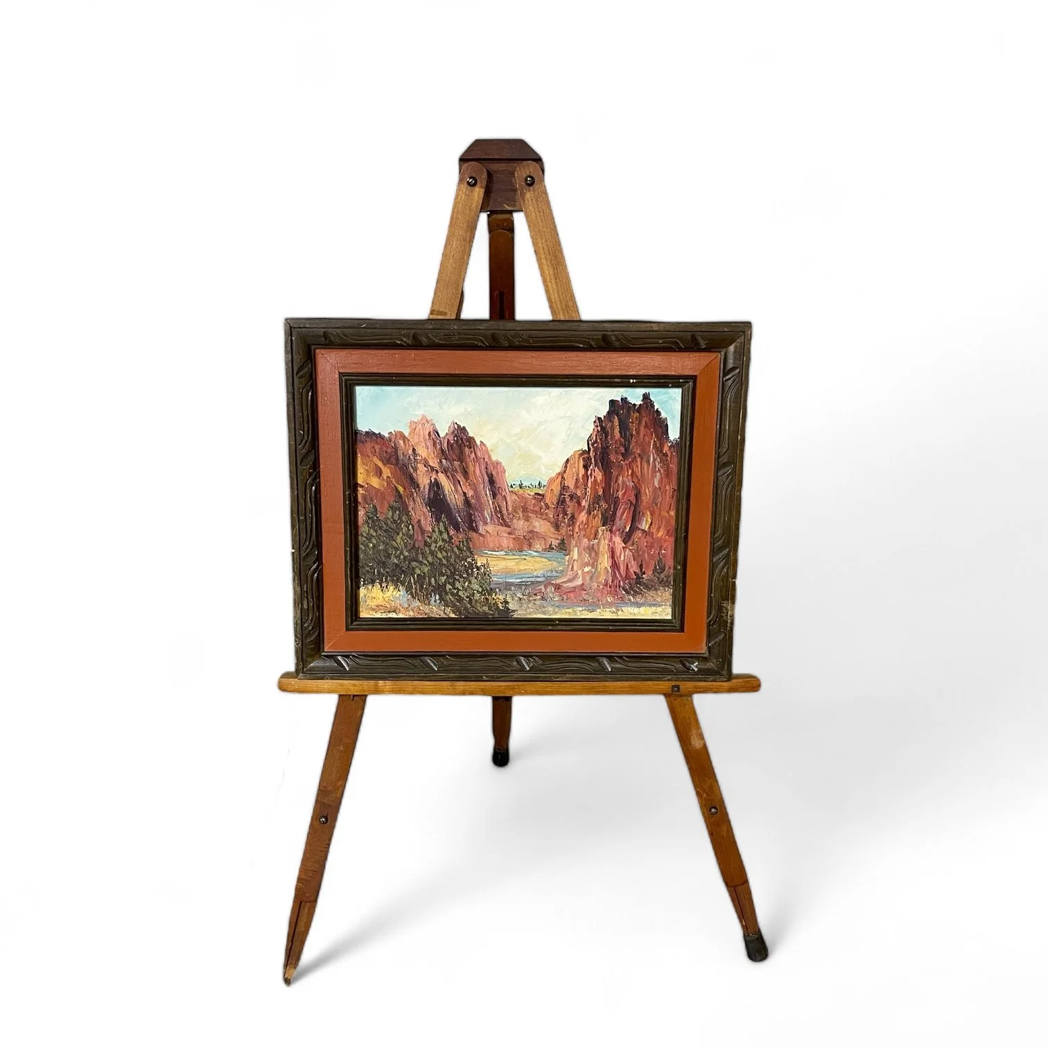 A vintage wooden adjustable floor easel displaying a framed and matted vintage oil painting of a rock canyon with a river, trees, and shrubs.