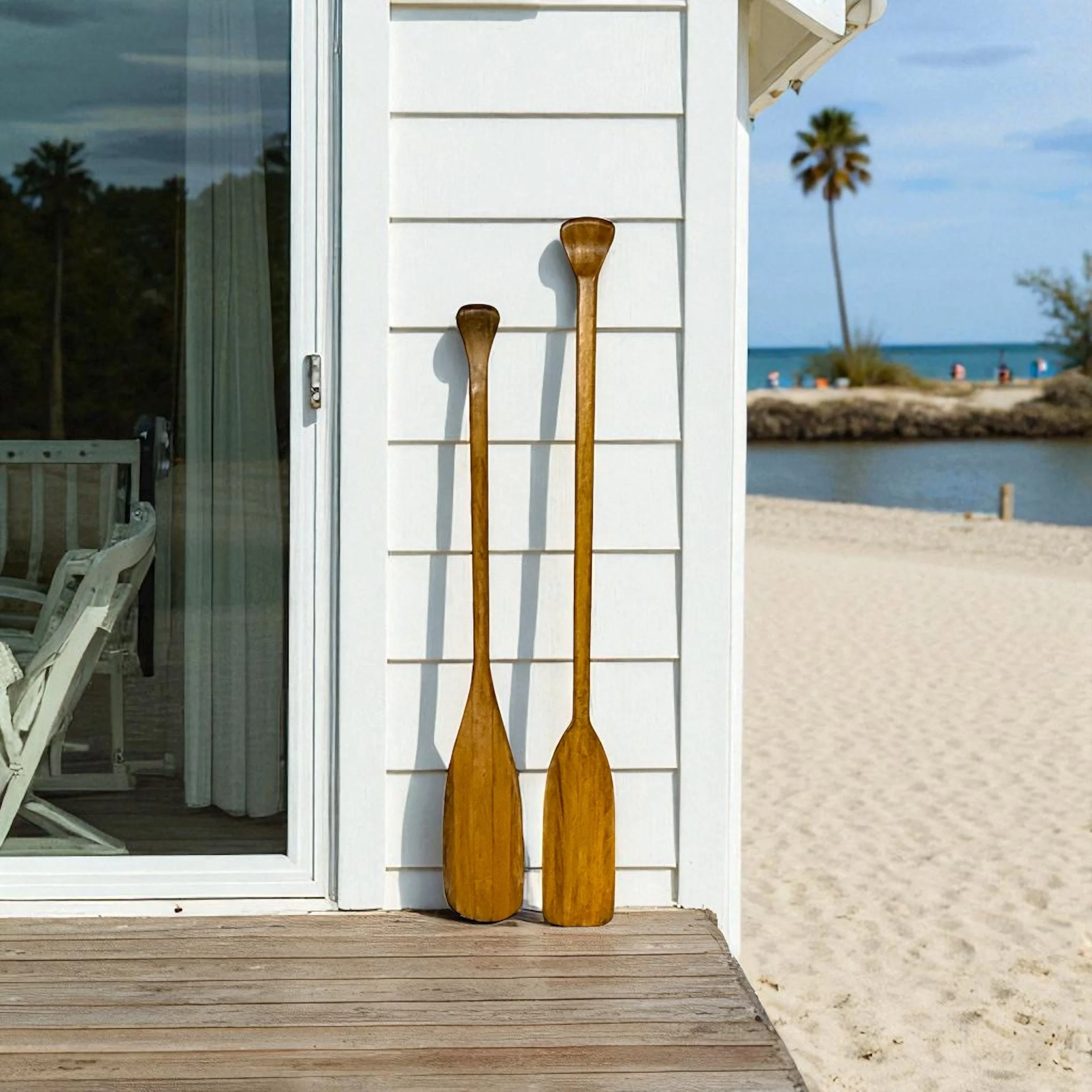 Vintage pair wooden boat oar paddles leaning against a white beach house with a sandy beach, water, and palm trees in the background.