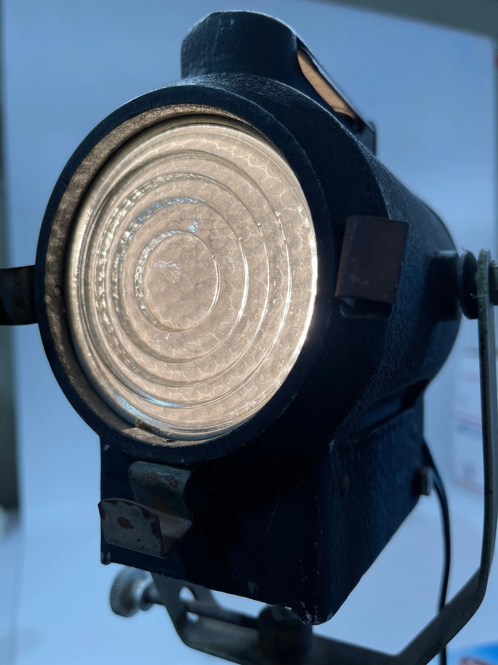 A vintage all original theater stage spotlight by Cycon, manufactured in Los Angeles, CA on a Acme Lite black metal adjustable tripod lamp in front of an industrial building.