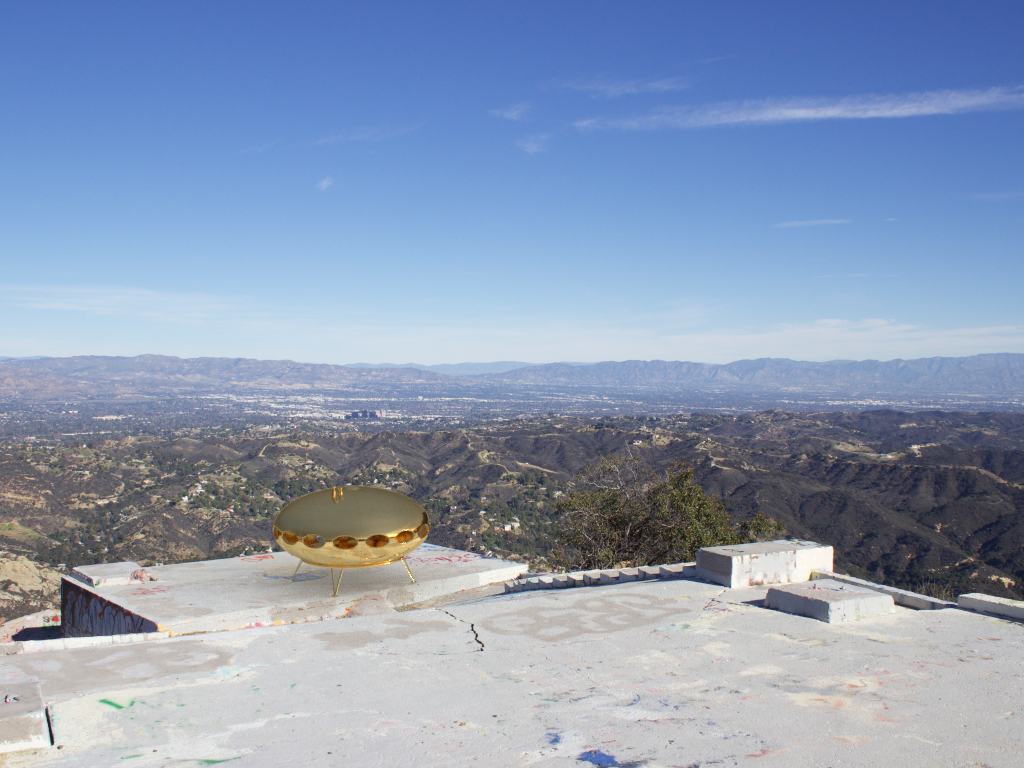  A photo of a gold Futuro Home which is an oval home designed to look like a spacecraft. The Futuro home sits on a concrete slab overlooking a canyon on high up mountain peek.  The photo was taken from a higher viewpoint up the mountain looking down 