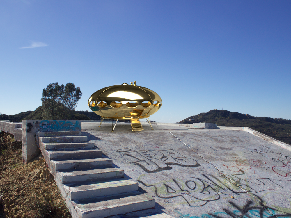  A photo of a gold Futuro Home which is an oval home designed to look like a spacecraft. The Futuro home sits on a concrete slab overlooking a canyon on high up mountain peek.   This photo is taken looking up at the Futuro home form a lower point on 