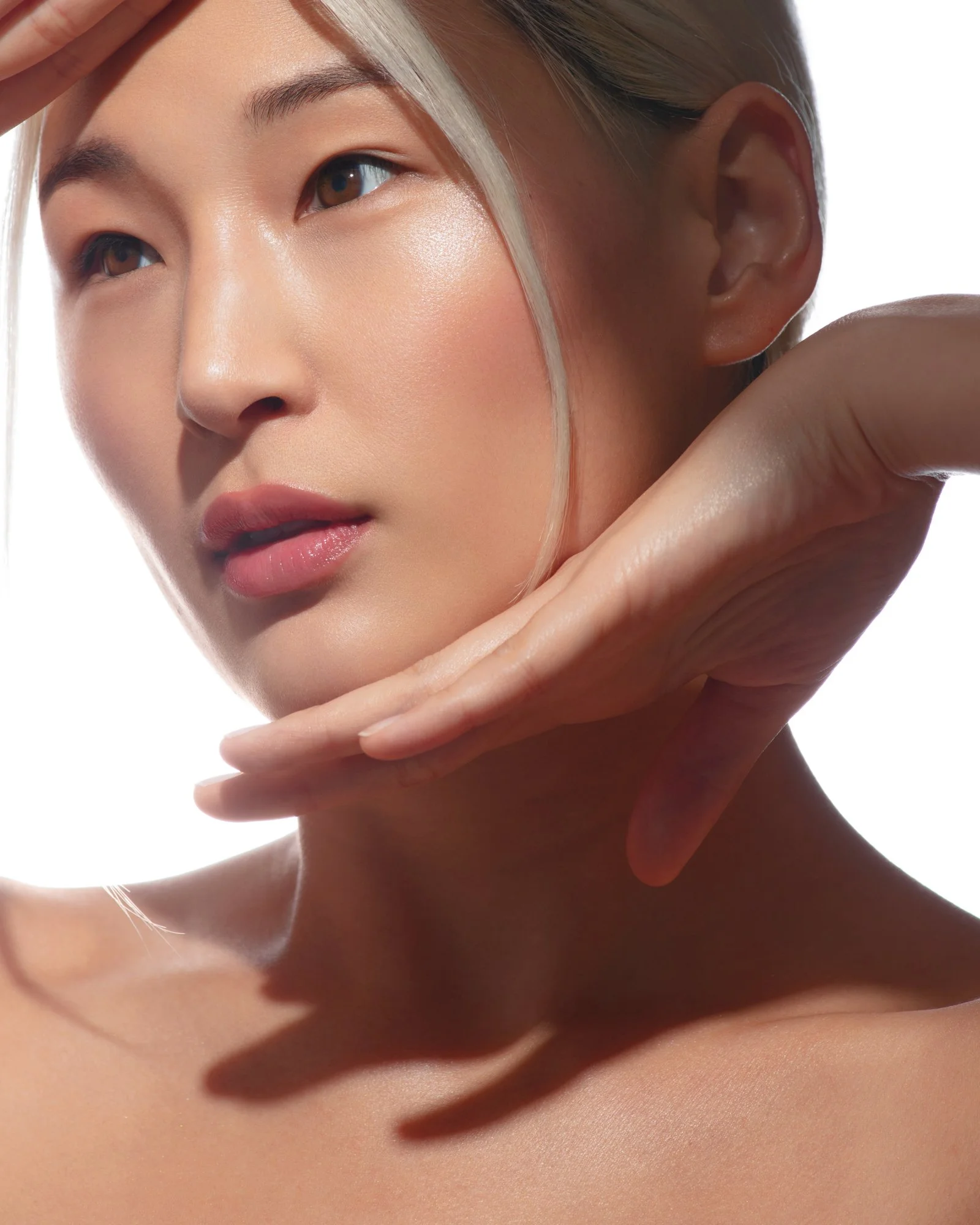 Close-up of a woman with flawless skin, slight makeup, and blonde hair, posing with her hand near her face against a plain white background.