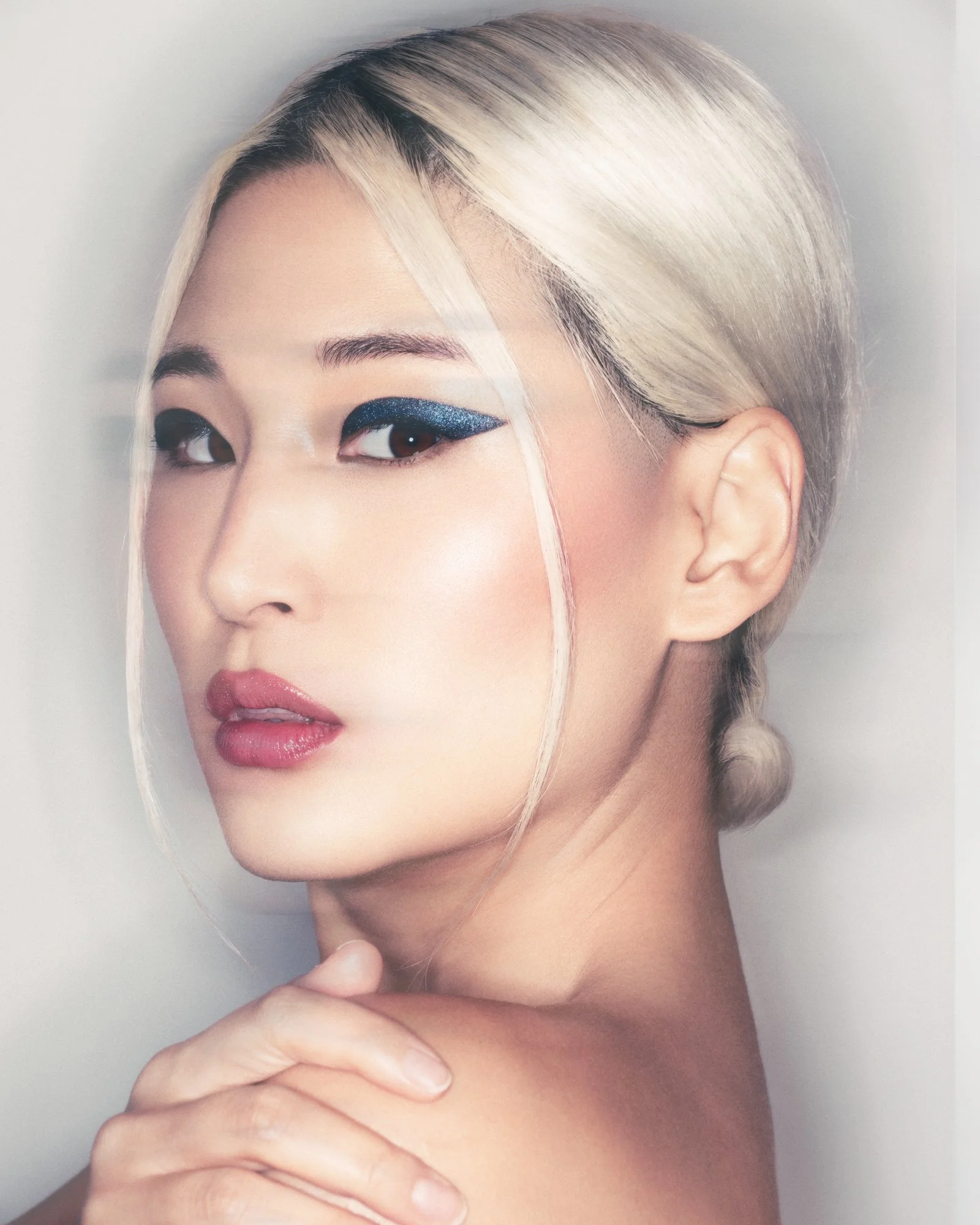 Close-up of a woman with platinum blonde hair, wearing blue glitter eyeshadow, light pink lipstick, and holding her hand near her chin against a neutral background.