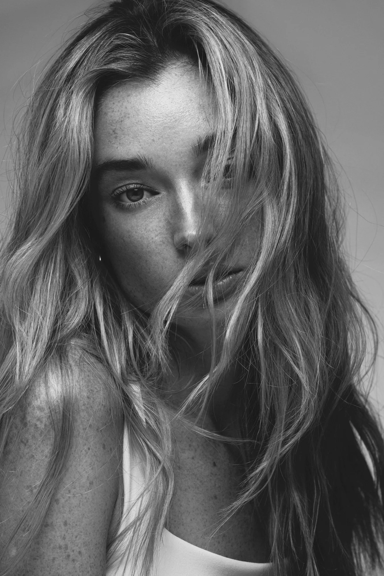 Close-up black and white photograph of a woman with wavy hair partially covering her face, showing one eye, freckles, and part of her lips.