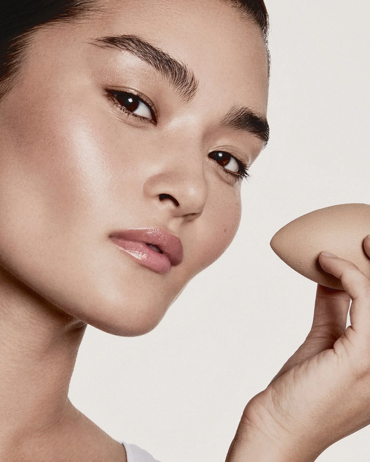 Close-up of a woman holding a makeup sponge near her face, with flawless skin, natural makeup, and groomed eyebrows, against a plain light background.