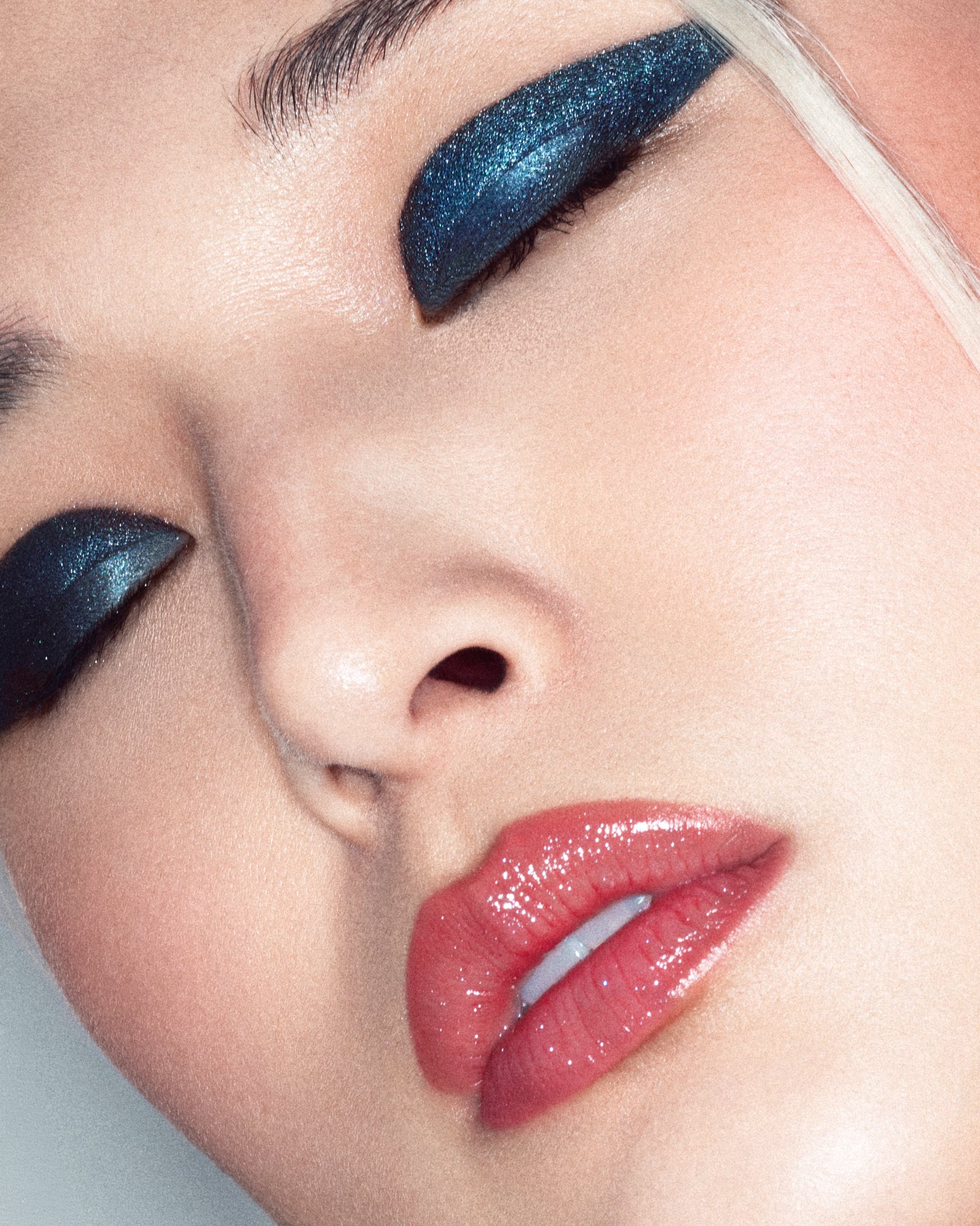 Close-up of a woman's face with vibrant blue and black glittery eyeshadow, glossy pink lips, and well-groomed eyebrows.