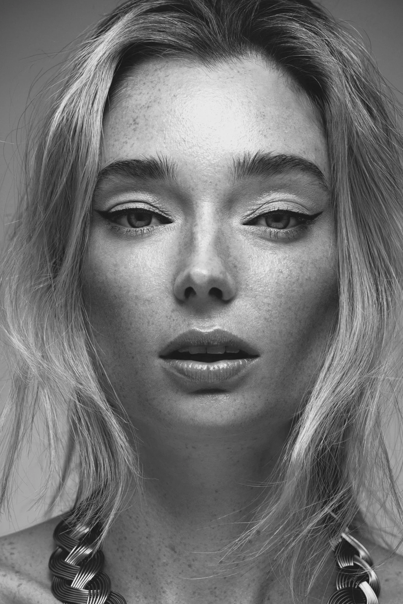 Black and white close-up portrait of a woman with long hair, textured skin, and makeup, wearing a beaded necklace.