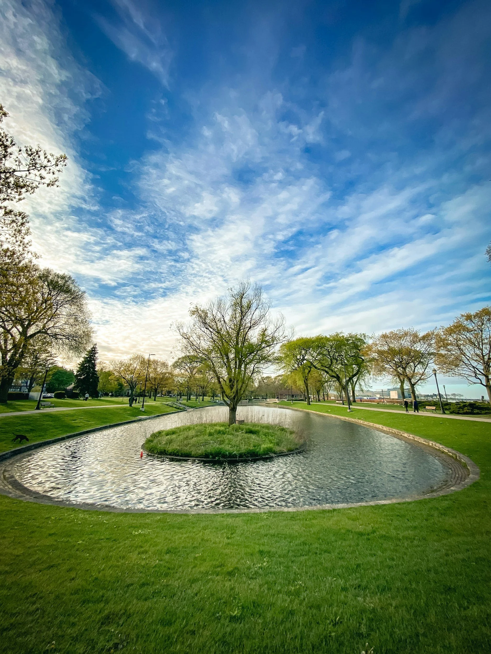Arrington Lagoon at Dawes Park with a small circular water feature at its center, surrounded by green grass and mature trees. The sky above is partly cloudy with patches of blue.