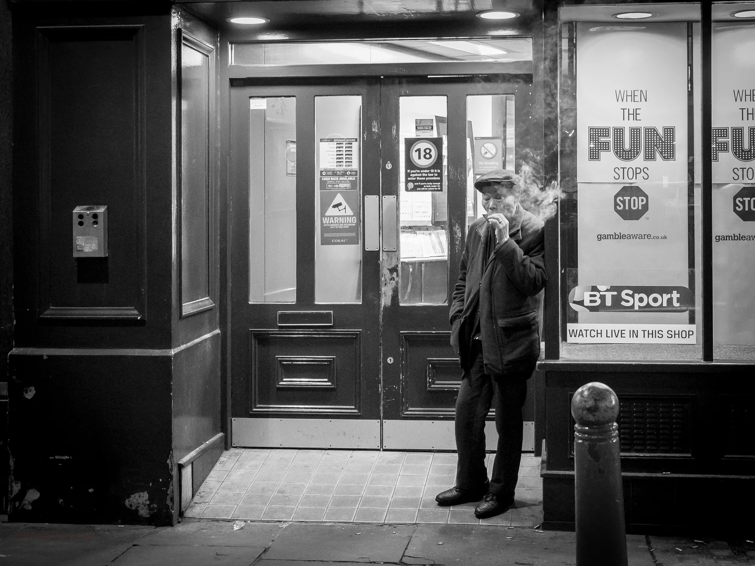 A dark scene featuring a man smoking (the smoke is obvious) leaning against the front of a betting shop (bookies as we say in the UK). The text reads 'When the Fun Stops, Stop'.Camera:&nbsp;Olympus OM-D E-M1.&nbsp;Lens:&nbsp;12-40mm f/2.8 PRO @&nbsp…