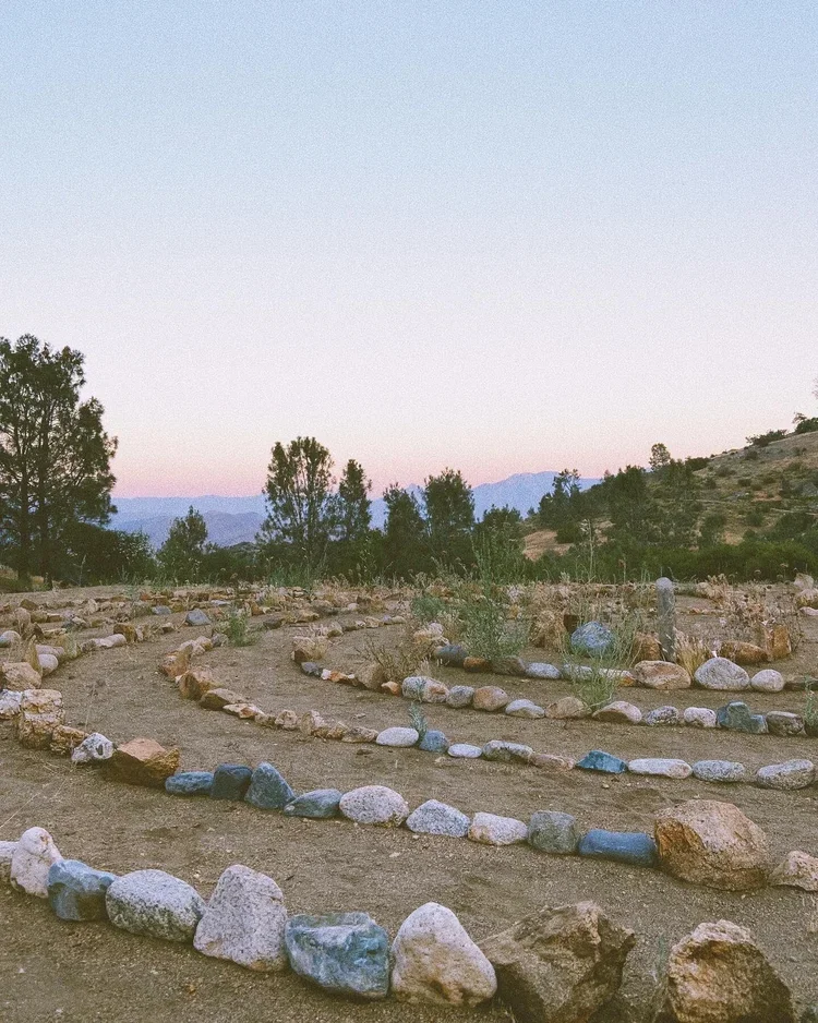 Rows of rocks forming a spiral pattern on a dirt ground outdoors, with bushes and trees in the background during dusk or dawn.