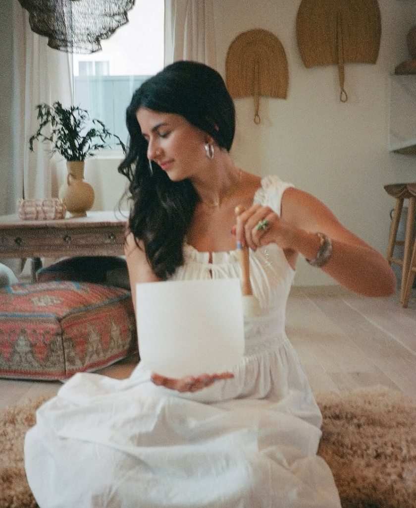 A woman with long dark hair in a white dress is seated on the floor, slicing a cake with a knife. The room has a rustic decor with a patterned cushion, wooden wall hangings, and a vase with greenery on a table.